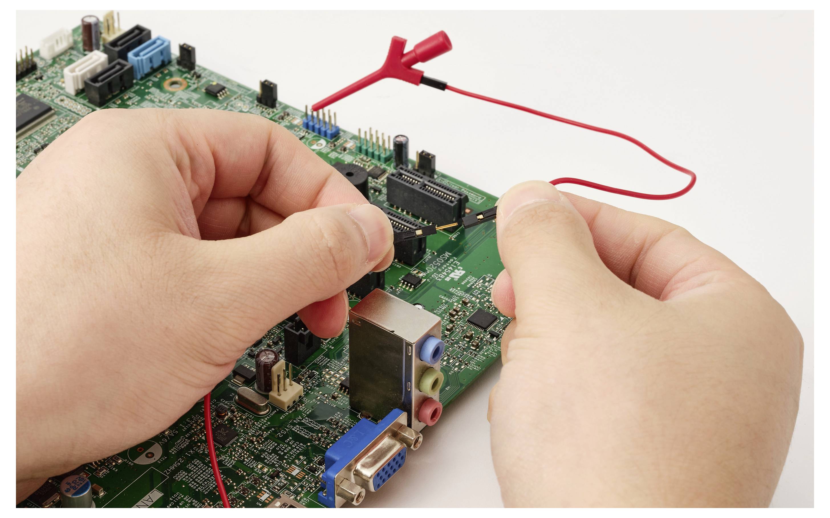 Handen gebruiken rode testdraden om een computermoederbord te onderzoeken, wat suggereert dat er elektronische troubleshooting of reparatie plaatsvindt.