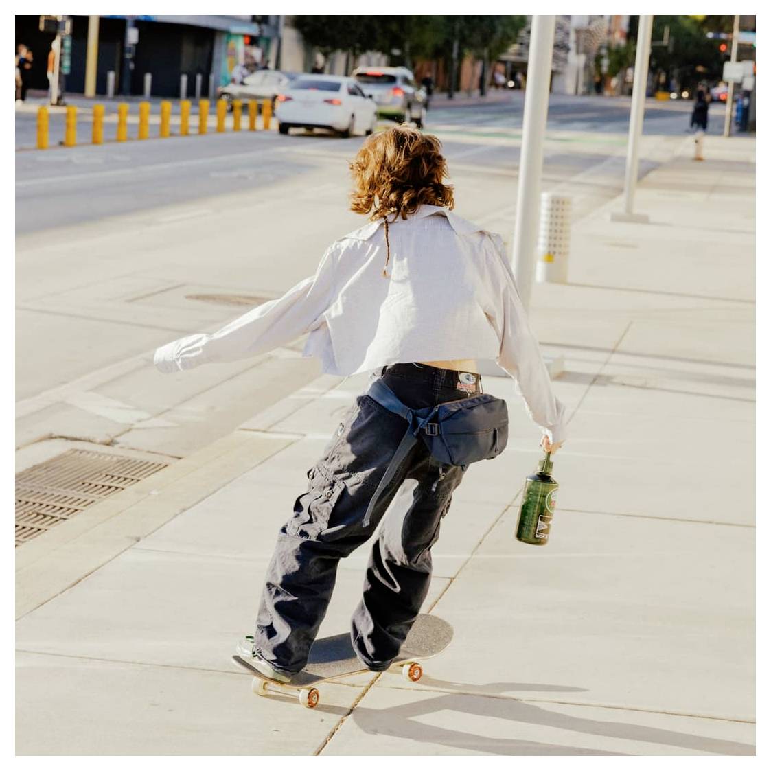 Persoon aan het skateboarden op een zonnig stadspad, met een groene fles in de hand, met auto's en straatpaaltjes op de achtergrond.