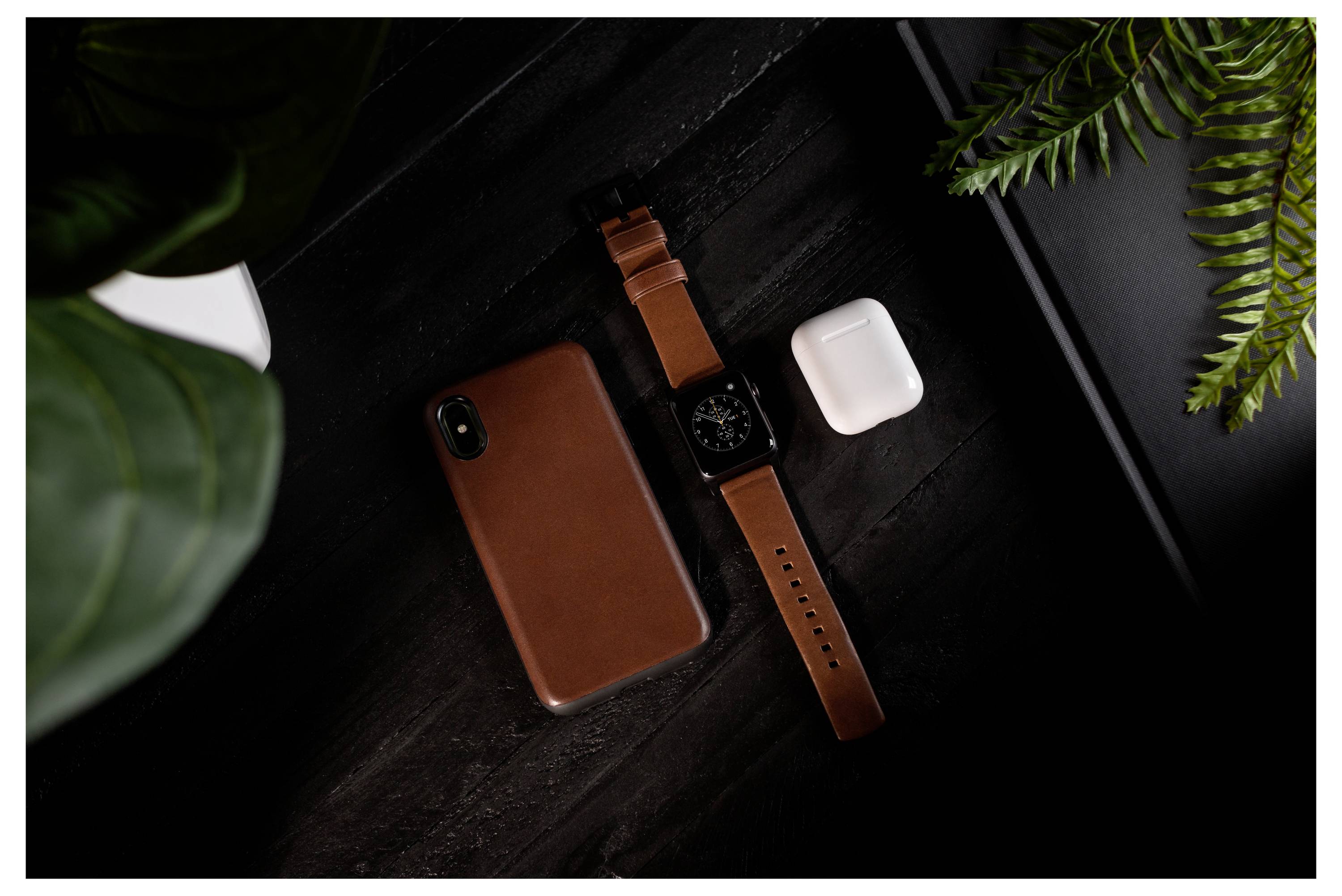 Bovenaanzicht van een bruine smartphone, smartwatch met lederen band en wit draadloze oordopjeshoesje op een donker bureau met plantenbladen.