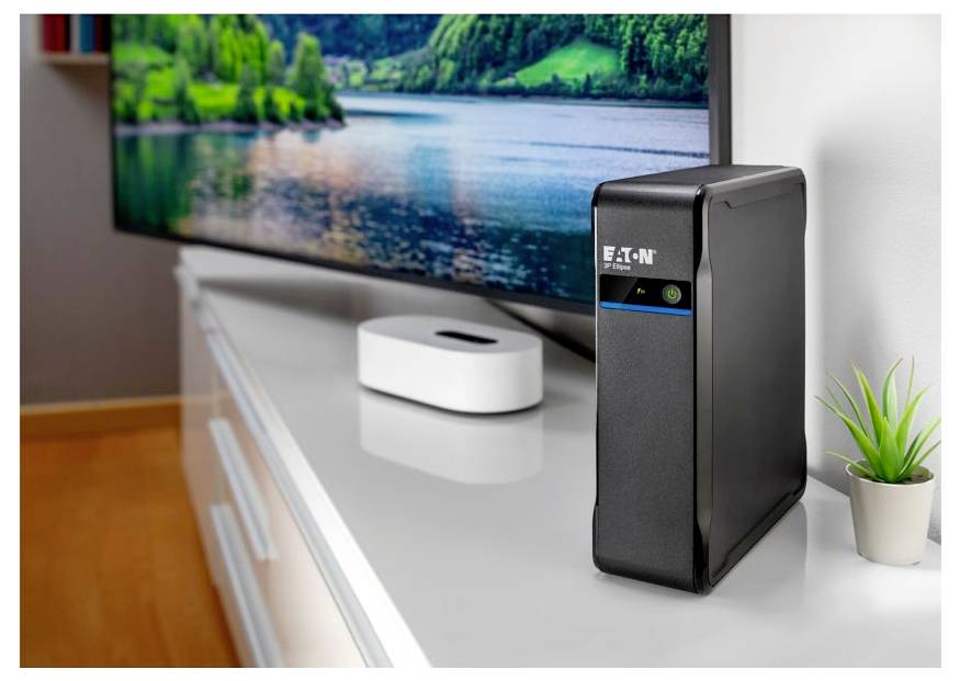 Een zwarte Eaton UPS op een tv-meubel naast een klein wit apparaat en een potplant, met een landschapsafbeelding weergegeven op de tv.