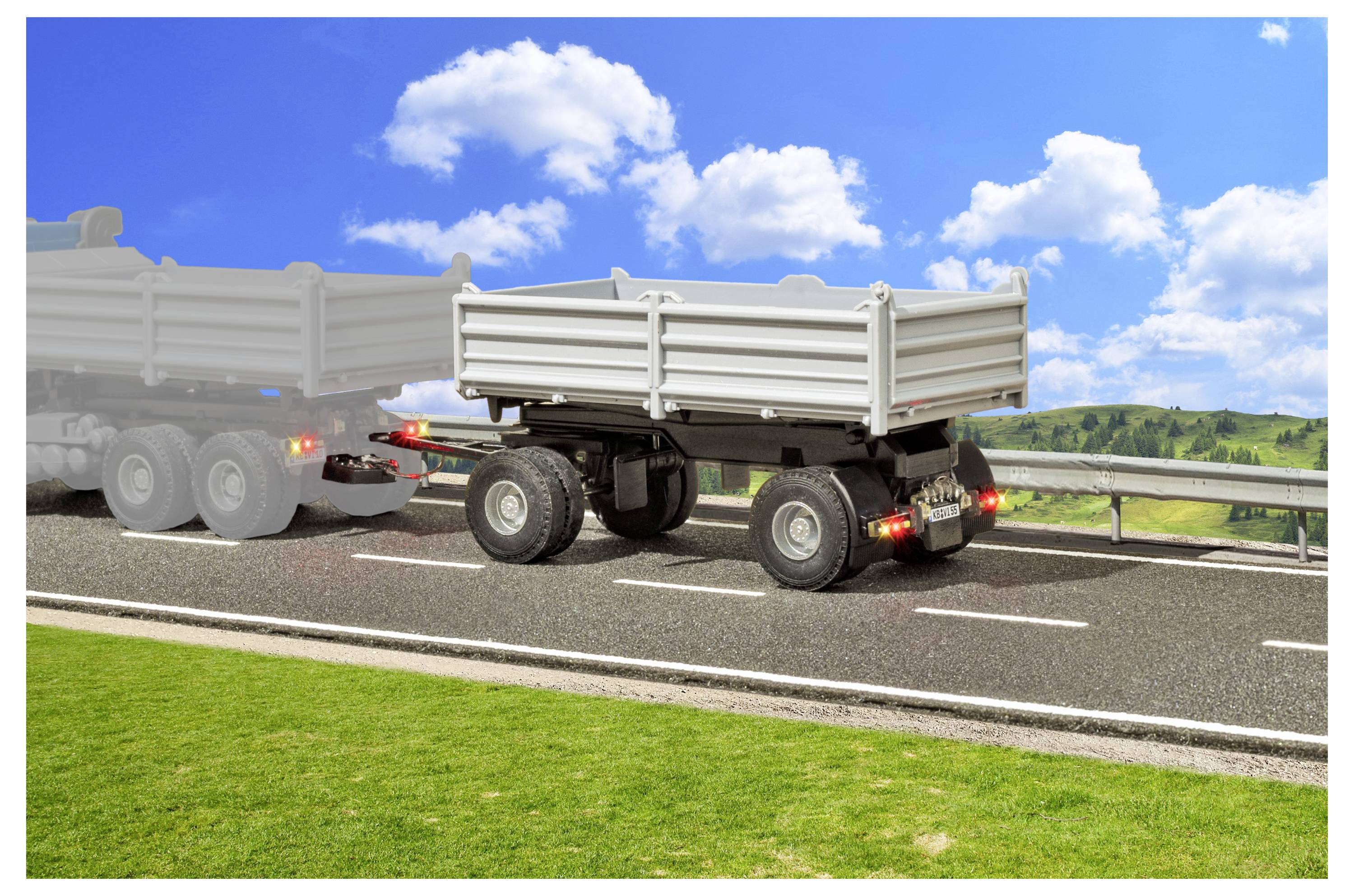 Zijkant van een grijze kiepwagen die door een vrachtwagen op een snelweg wordt getrokken, met wegversperringen, groen gras en een helderblauw lucht met wolken.