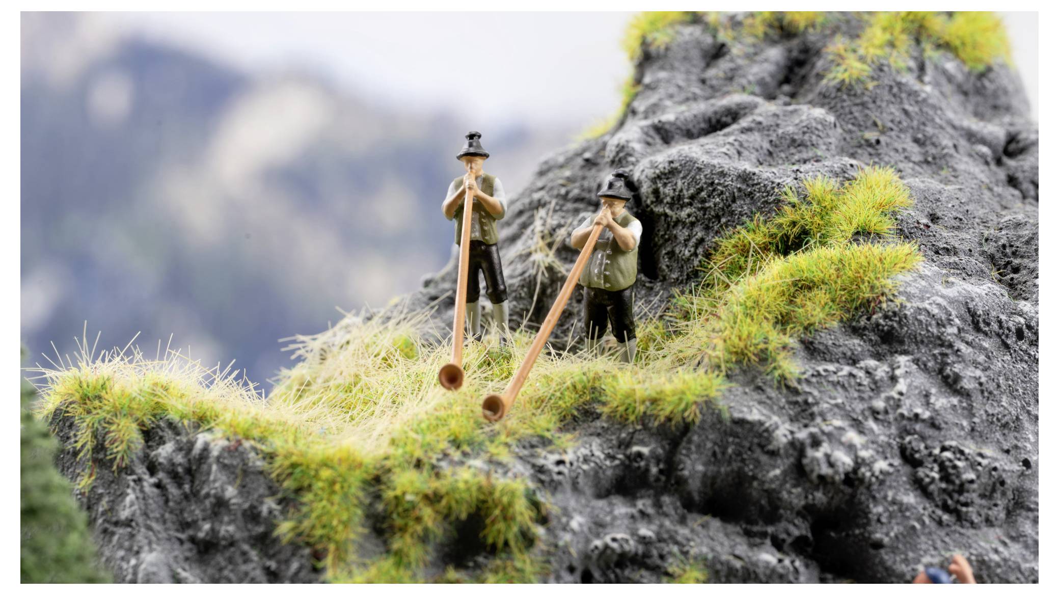Twee wandelaars spelen lange houten didgeridoo's op een rotsachtige bergrichel met gras, tegen een wazig alpien decor.