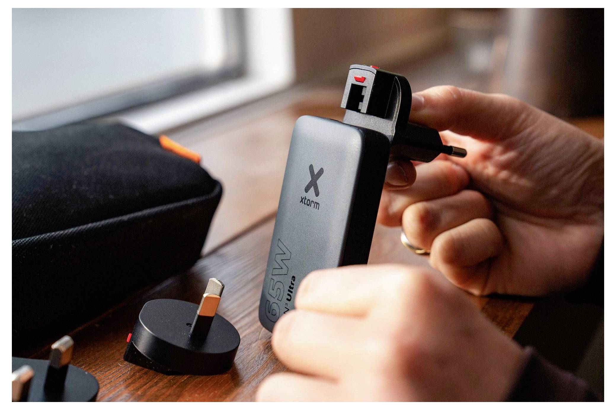 Handen die een Britse adapter in een Xiaomi draagbare powerbank op een tafel steken, met een oplaadkassette en andere stekkers in de buurt.