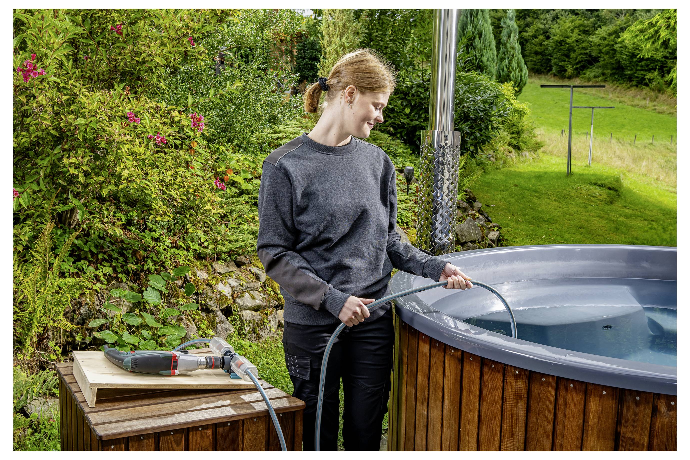 Vrouw die een houten hottub buiten met een slang vult naast een tafel met gereedschap in een tuin.