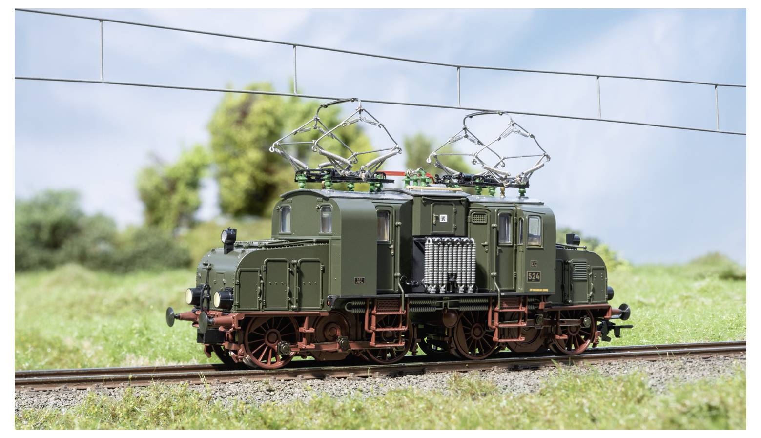 Een groen modelelectrische locomotief op rails, met verheven stroombakken onder bovenleiding in een groen buitenterrein.
