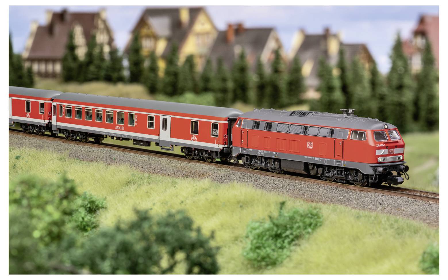 Rode modeltrein die rode passagierswagons langs een gebogen spoor trekt naast grasachtig landschap, met onscherpe dorpshuizen op de achtergrond.