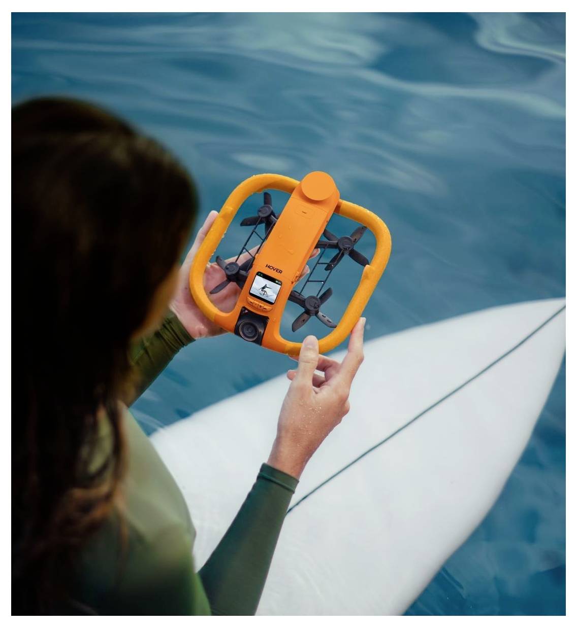 Een persoon die een oranje drone boven helder blauw water vasthoudt naast een wit surfboard, zich voorbereidt om deze op te stijgen of te inspecteren.