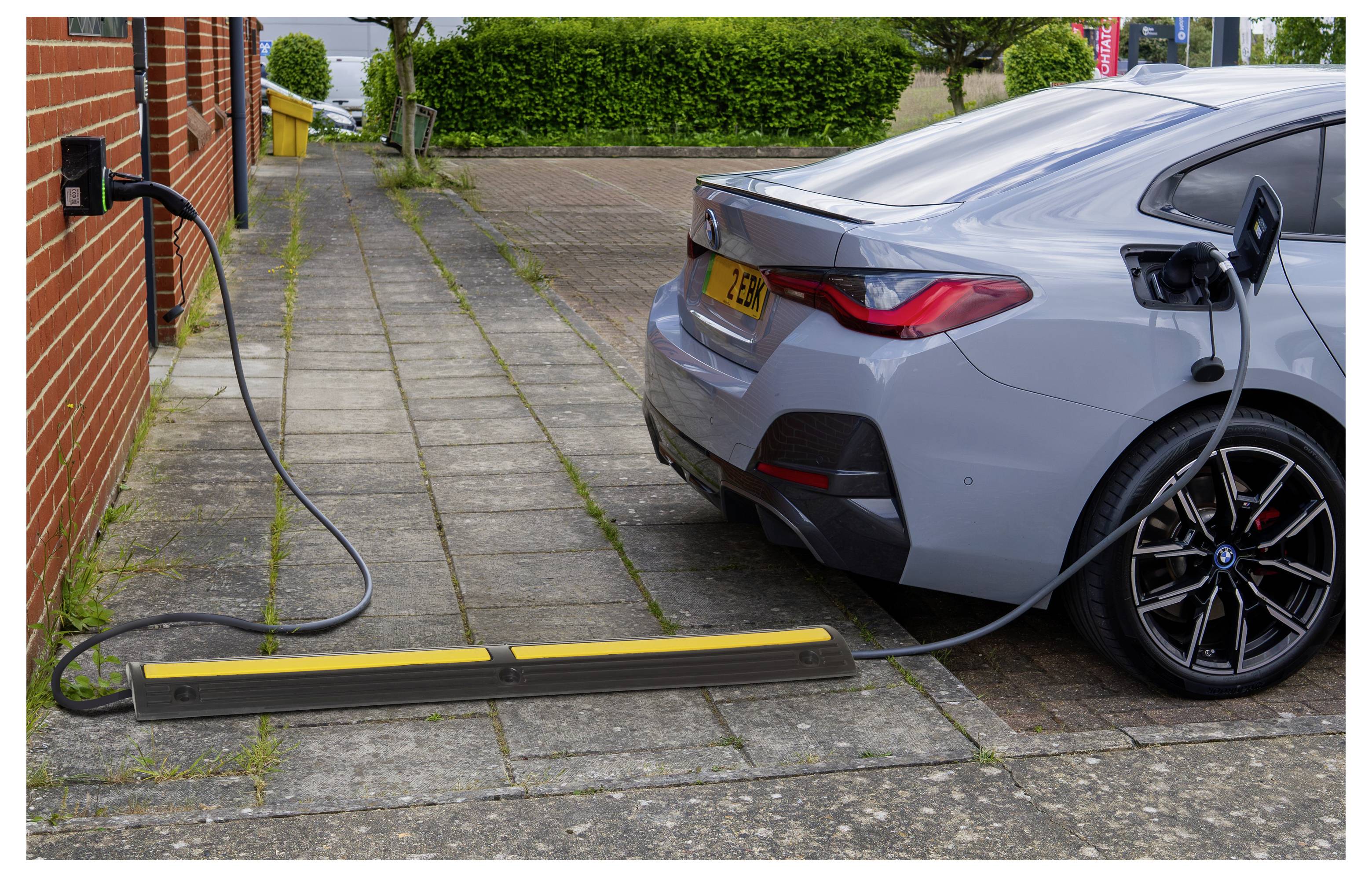 Grijze elektrische auto die wordt opgeladen vanaf een stopcontact aan de muur naast een bakstenen gebouw, met een kabel die over het trottoir loopt naar een op de oprit gemonteerd laadplatform.