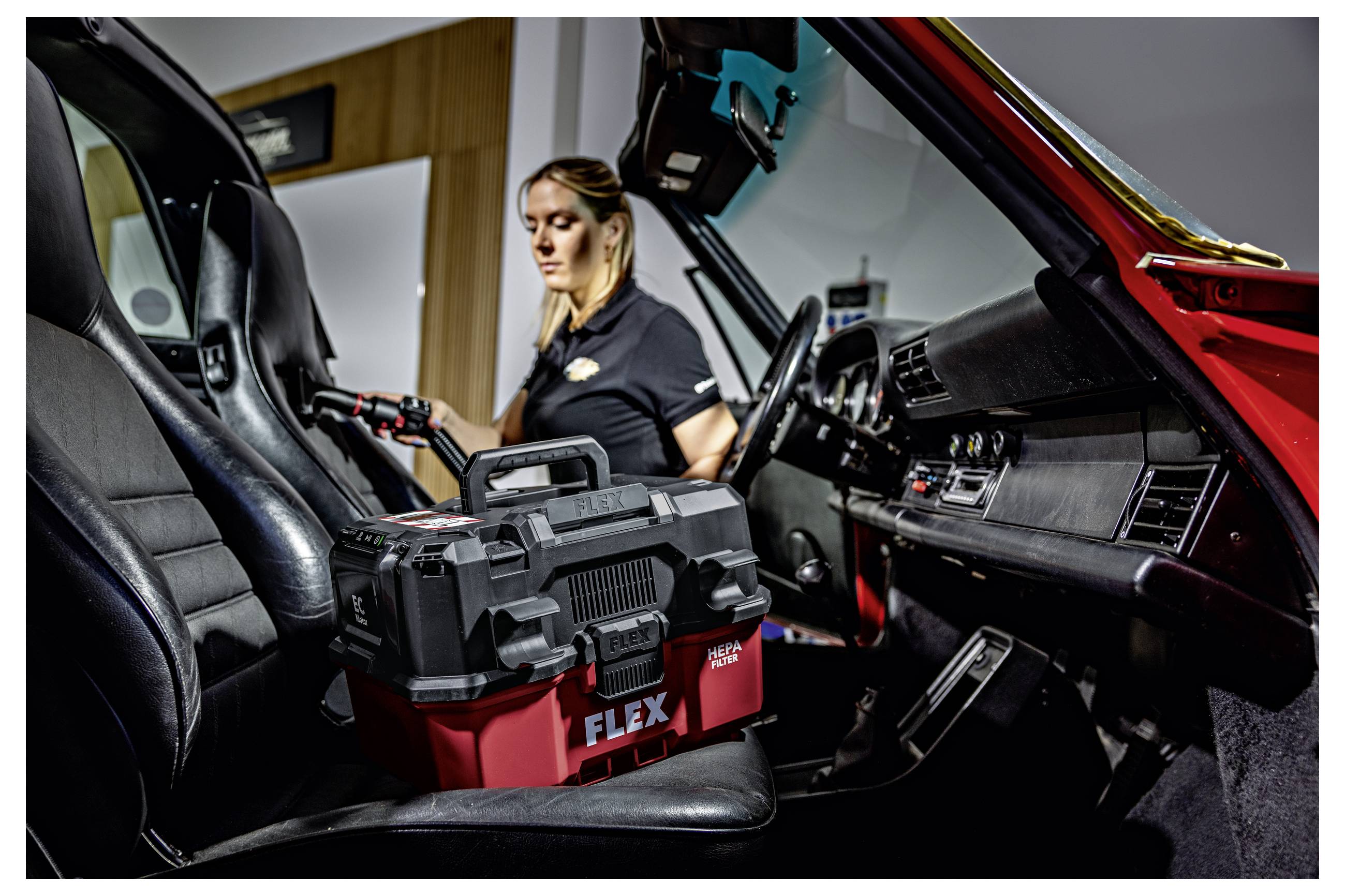 Vrouw in een auto-interieur die het schoonmaakt of detailt met een FLEX-stofzuiger/machine op de voorstoelen, met het dashboard en rode deur zichtbaar.