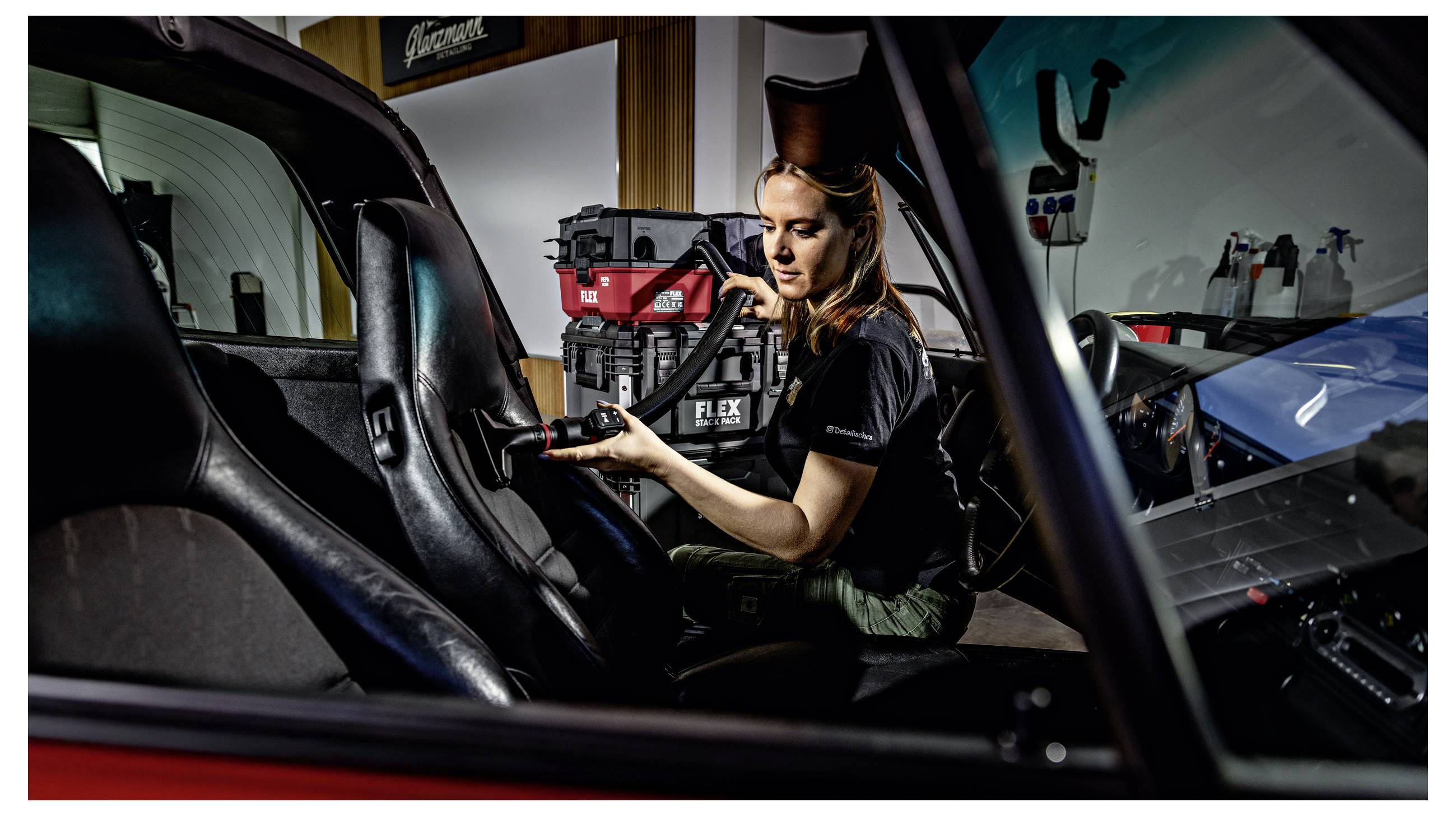 Vrouw die een rood en zwart FLEX elektrisch gereedschap bedient in een voertuigwerkplaats, zittend in een auto-interieur terwijl zij reparatie- of onderhoudswerkzaamheden aan apparatuur uitvoert.