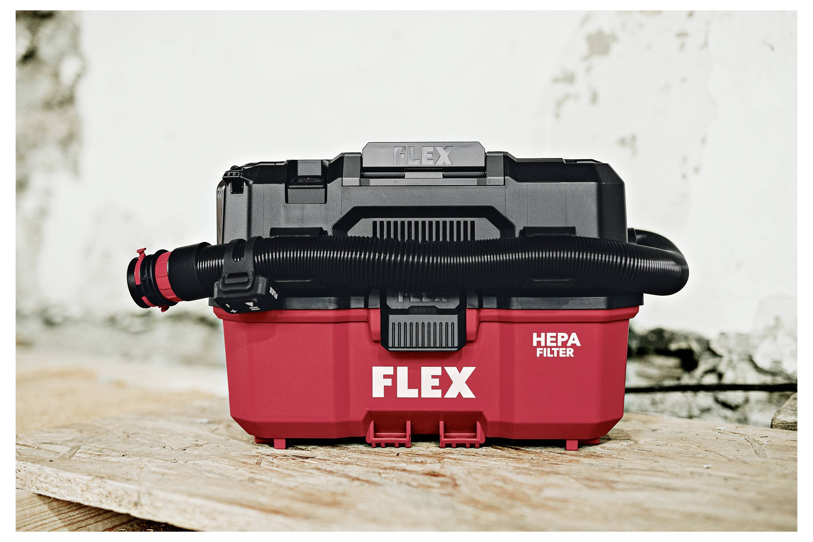 Rode FLEX HEPA-filterstofzuiger met zwarte slang eraan bevestigd, staande op een houten oppervlak tegen een lichte achtergrond.