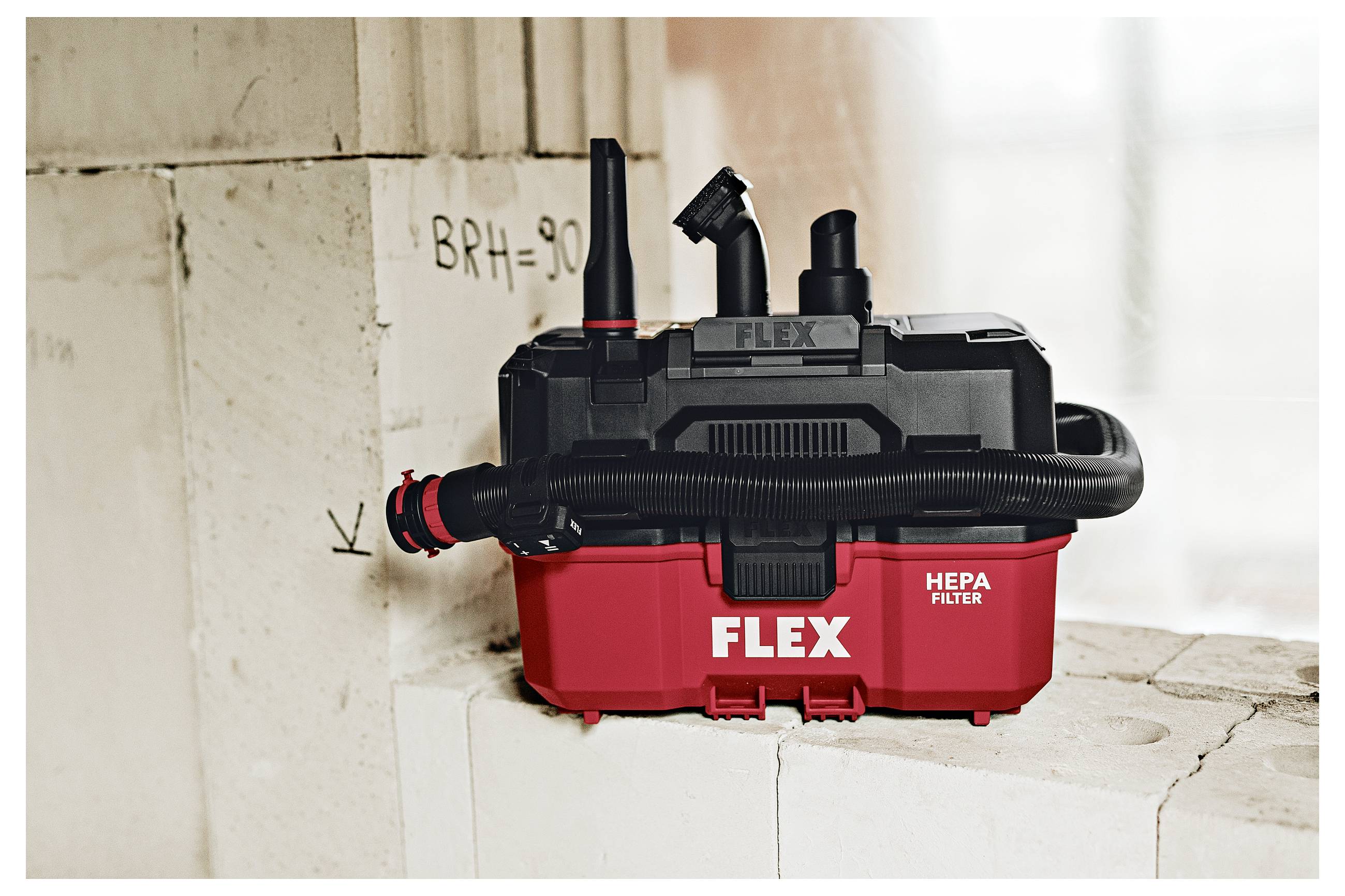 Rode FLEX HEPA-filterslofzuiger aan de muur gemonteerd, met slang en hulpstukken zichtbaar in een bouwomgeving.