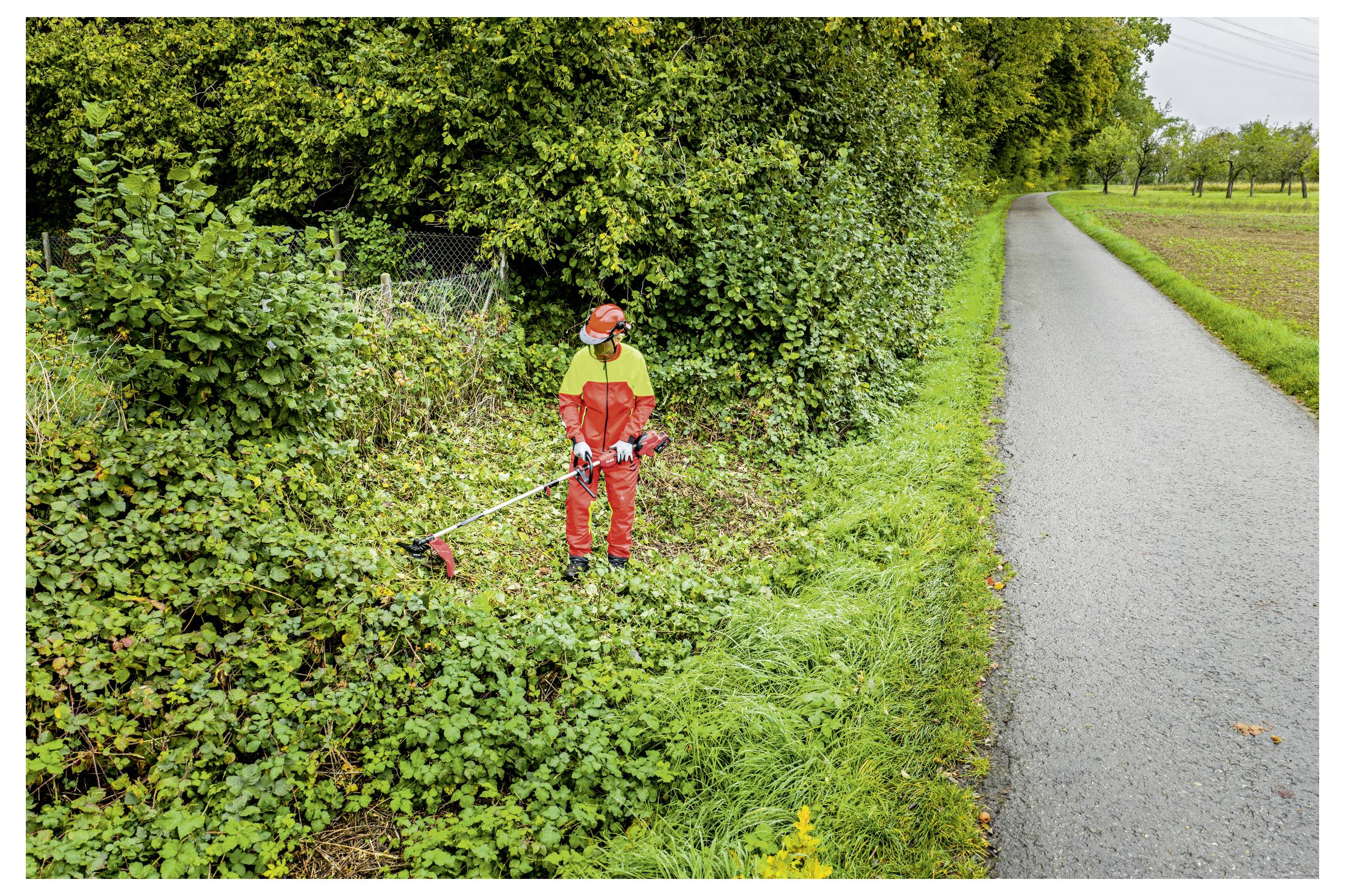 Een werknemer in oranje veiligheidskleding gebruikt een elektrische grasmaaier om overwoekerde wegkantvegetatie naast een landelijk geasfalteerde weg af te snijden.