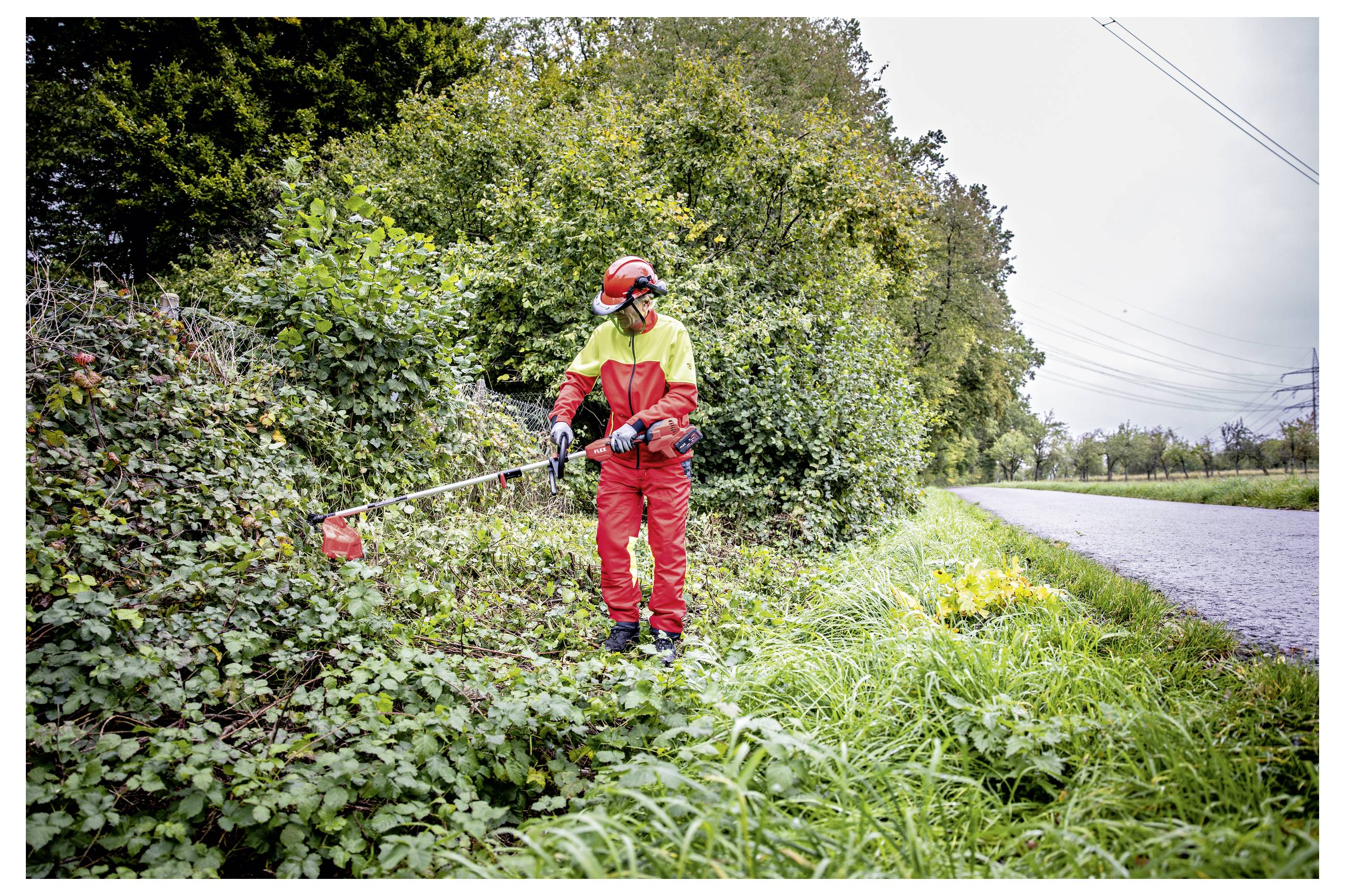 Werknemer in rode beschermende uitrusting die langs een plattelandsweg plantegroeien aan de wegkant bijsnijdt met een draaitrimmer.