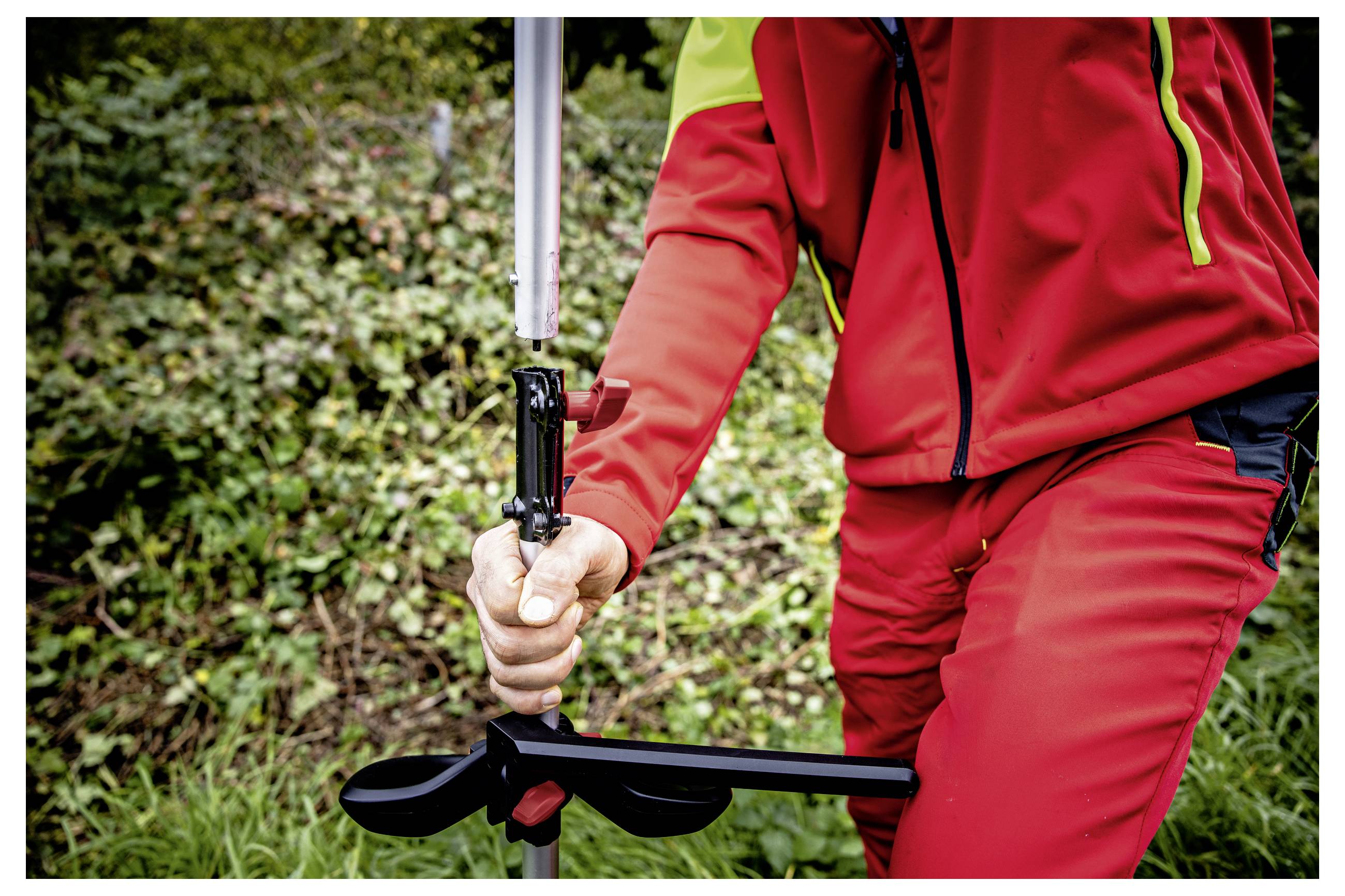 Close-up van een persoon in rode buitenkleding die een paal of gereedschap met een zwarte klem vasthoudt, buiten in de buurt van gras en struiken.