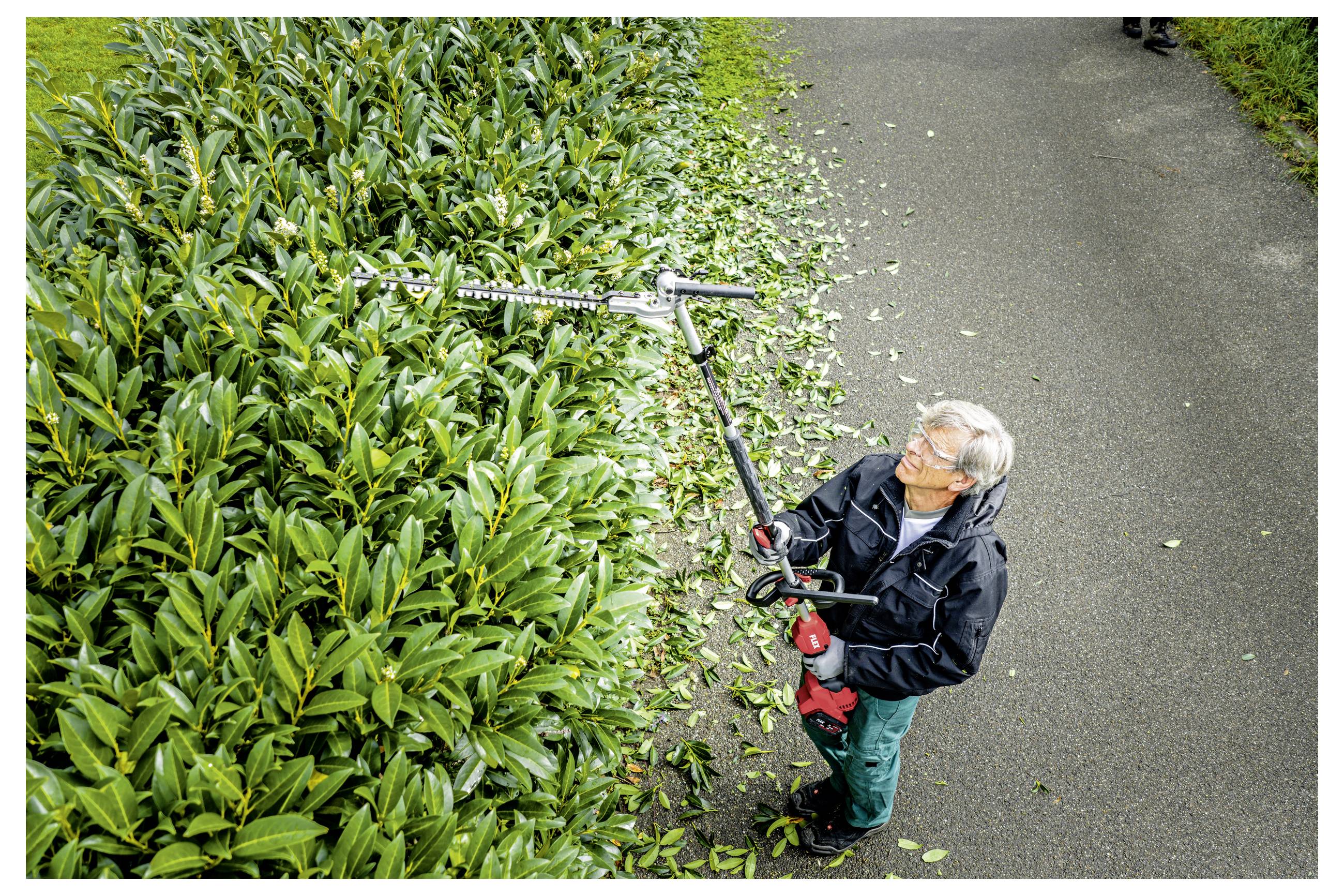 Een arbeider die een dichte haag bijsnijdt met een langstielige haagschaar naast een verharde weg, met afgesneden bladeren verspreid op de grond.