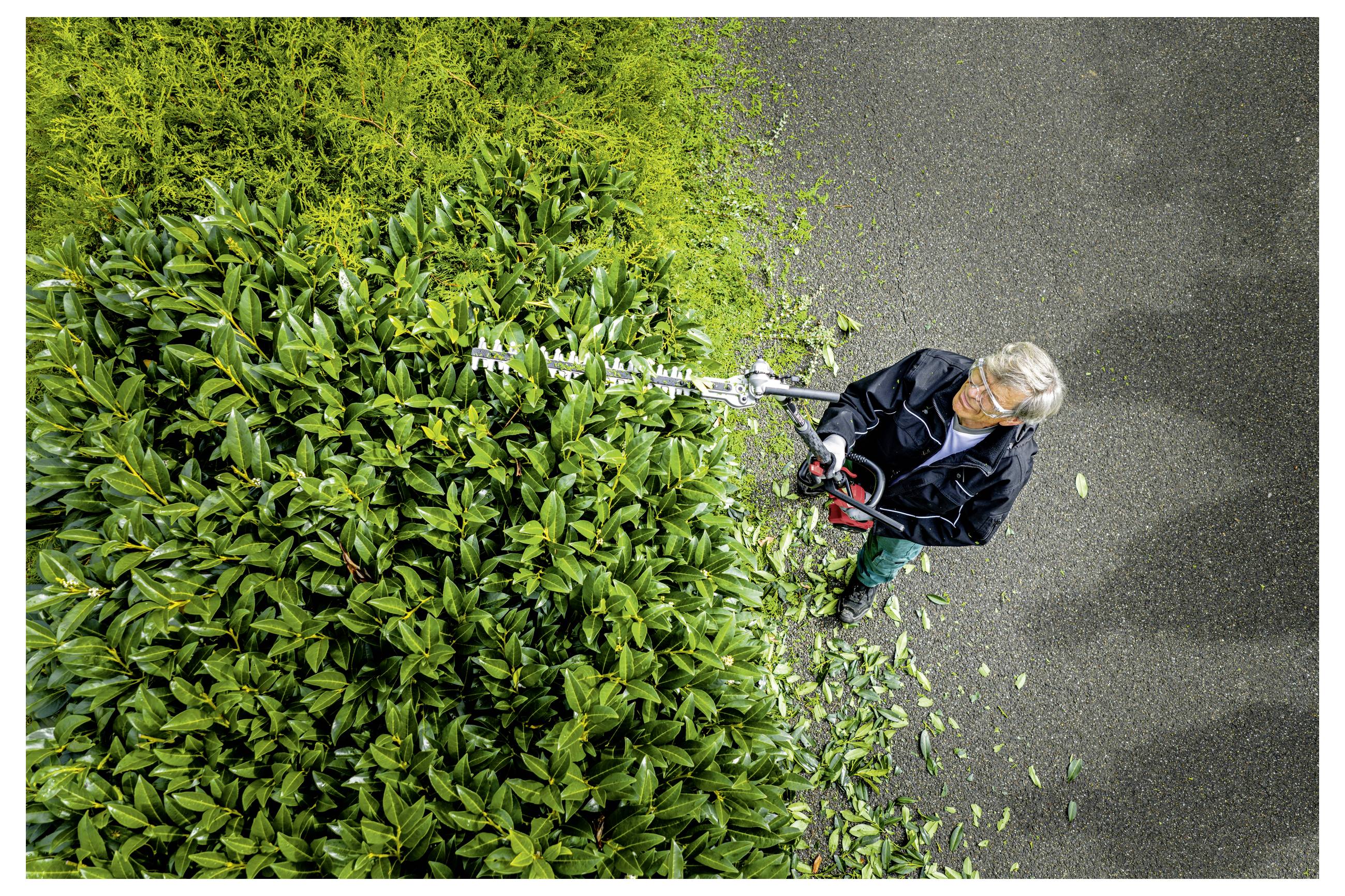 Bovenaanzicht van een werknemer die een grote groene haag snoeit met een lange haagschaar naast een geplaveid pad, met versneden bladeren verspreid op de grond.