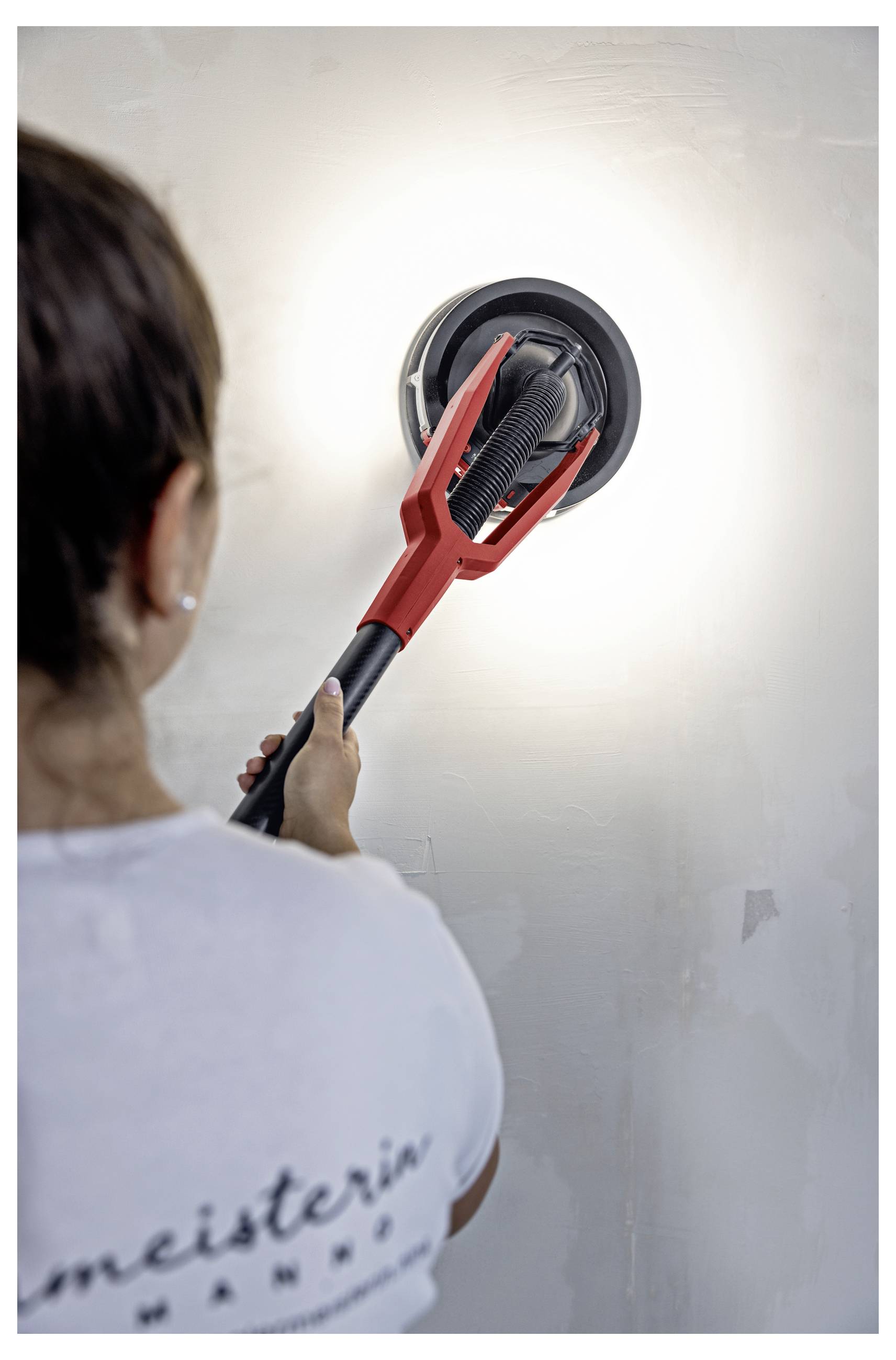 Een persoon die een muur met een rode drywallschuurmachine schuurt, waardoor het oppervlak wordt voorbereid voor schilderwerk of reparatie.