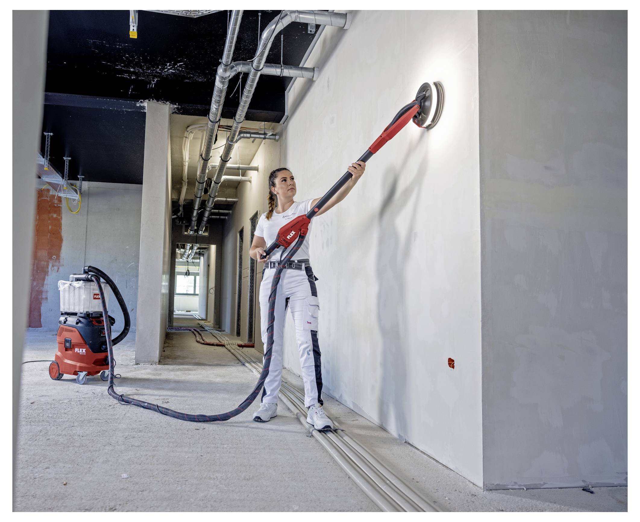 Arbeider die een lange muurschuurmachine gebruikt om een geplasterde muur in een onafgewerkte binnengangkamer glad te maken, met een vacuümunit op de grond.