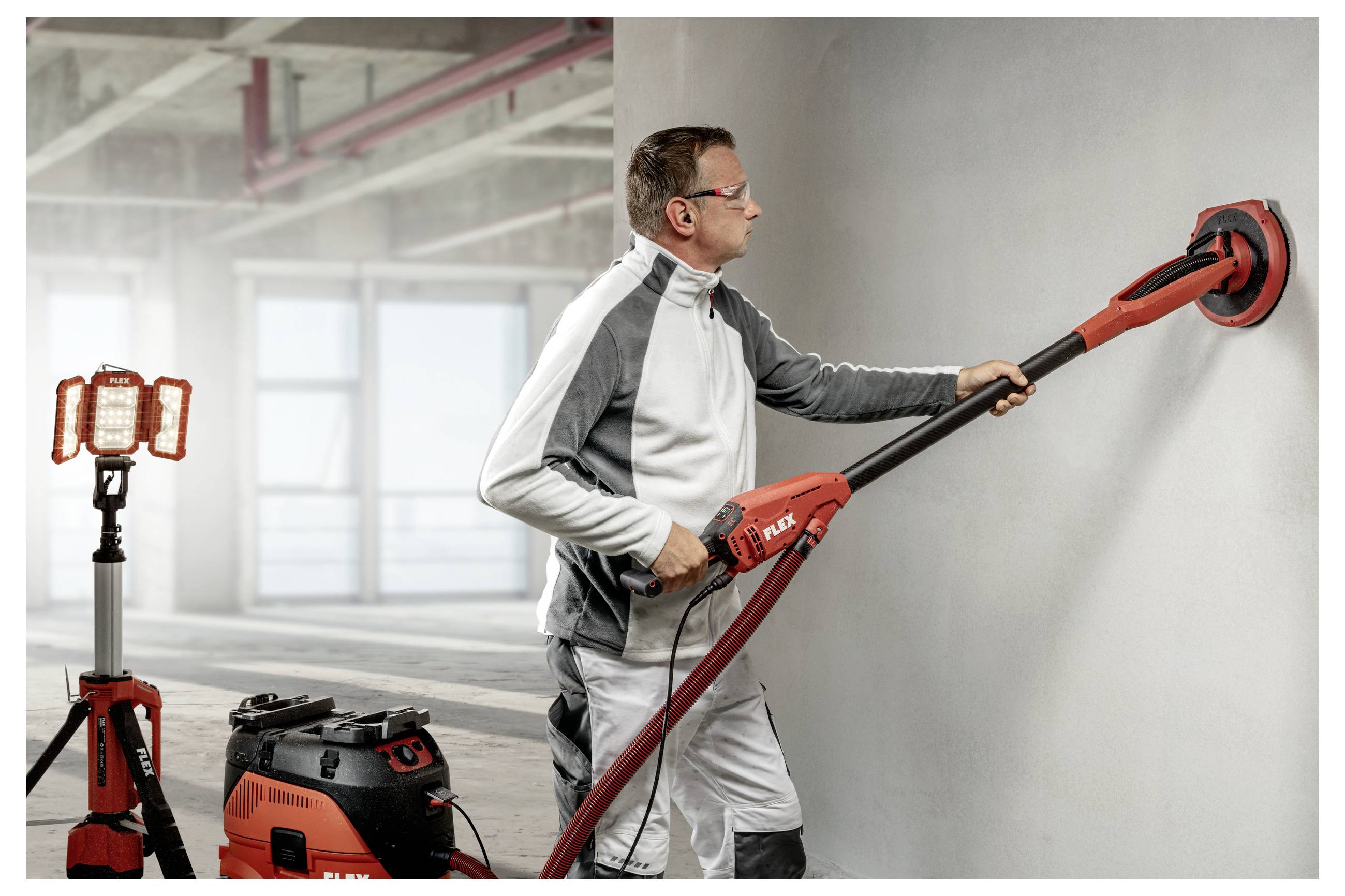Werknemer gebruikt een rood-zwart lang handvat wandschuurder op een grijze muur in een bouwruimte, met een draagbare werklamp en stofzuiger ernaast.