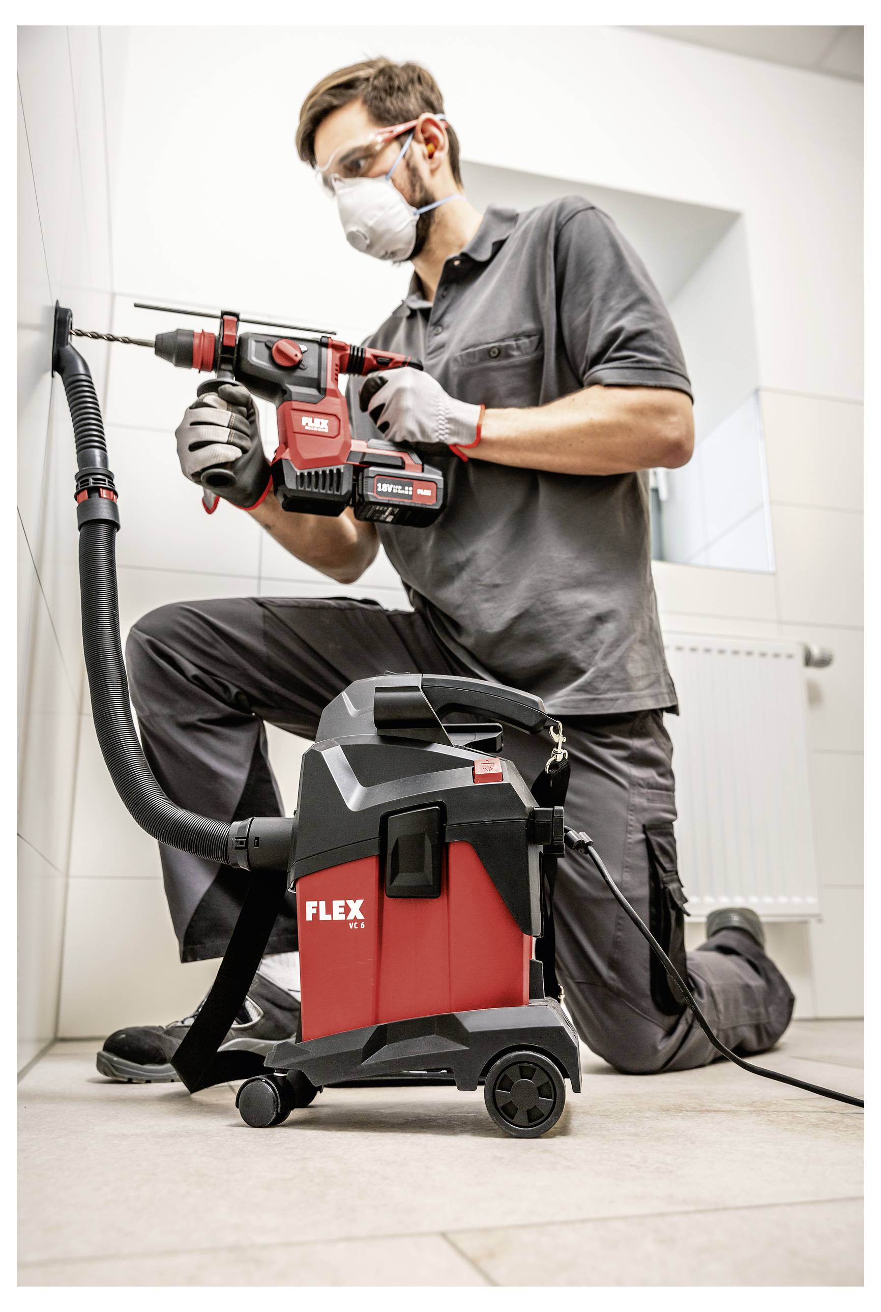 Werknemer met een masker die knielend binnenshuis met een rood FLEX-boormachine met stofafzuiger werkt.