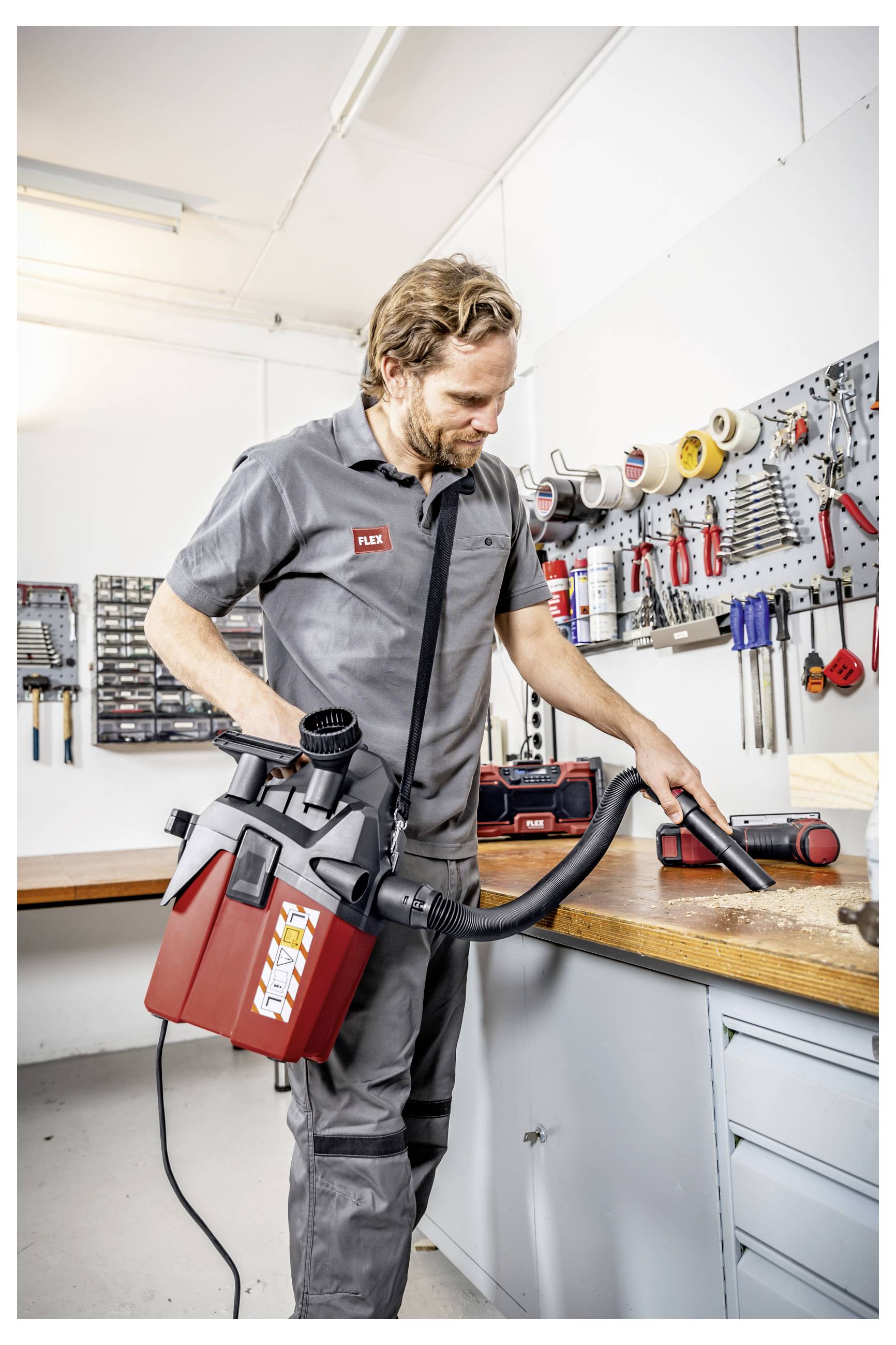 Monteur die een draagbare nat-droog stofzuiger gebruikt om een werkbank in een werkplaats schoon te maken, met gereedschap en benodigdheden georganiseerd op de muur achter hem.