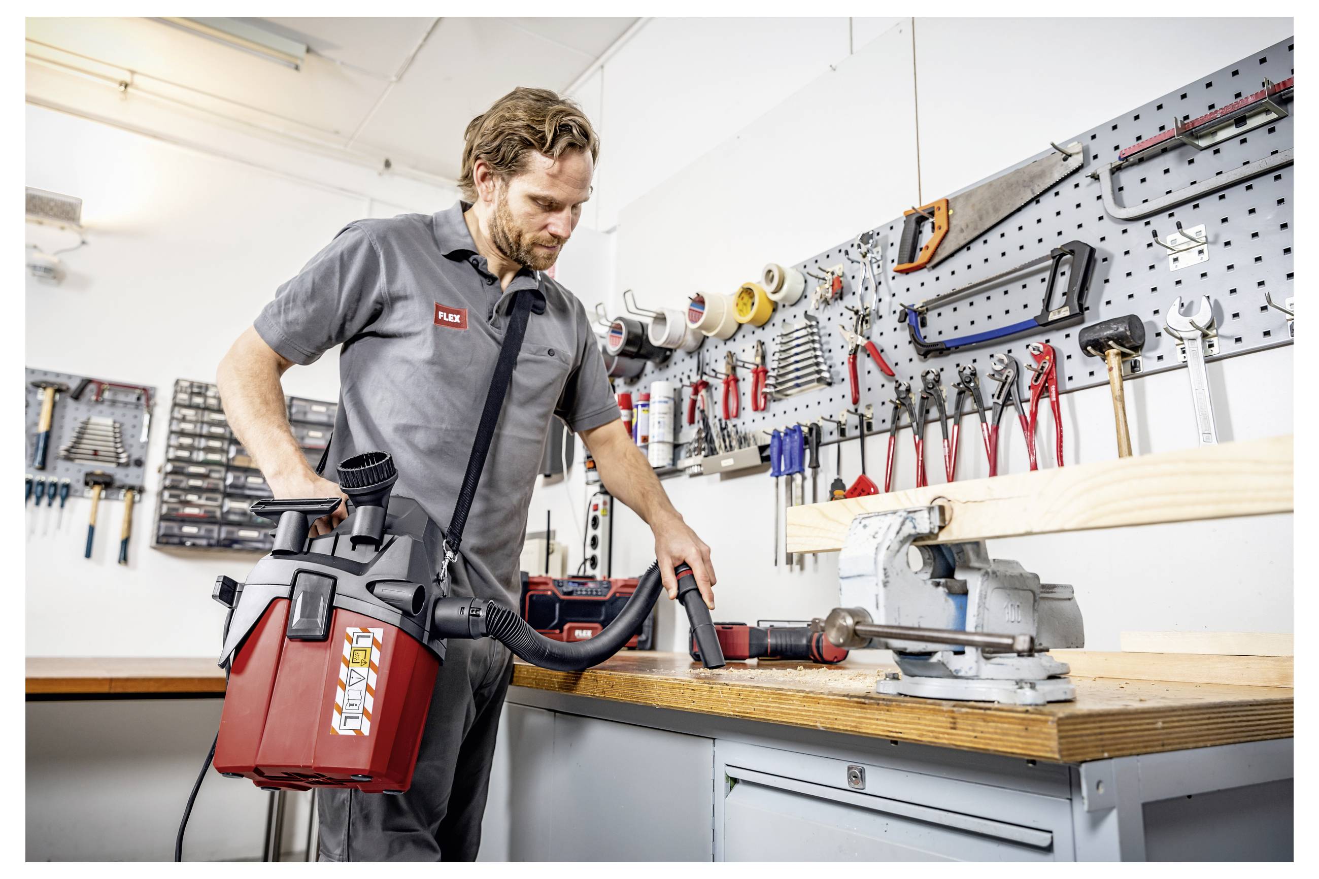 Een man in een werkplaats gebruikt een rode nat/droge stofzuiger om een werkbank schoon te maken naast een bankschroef en hangende handgereedschappen.