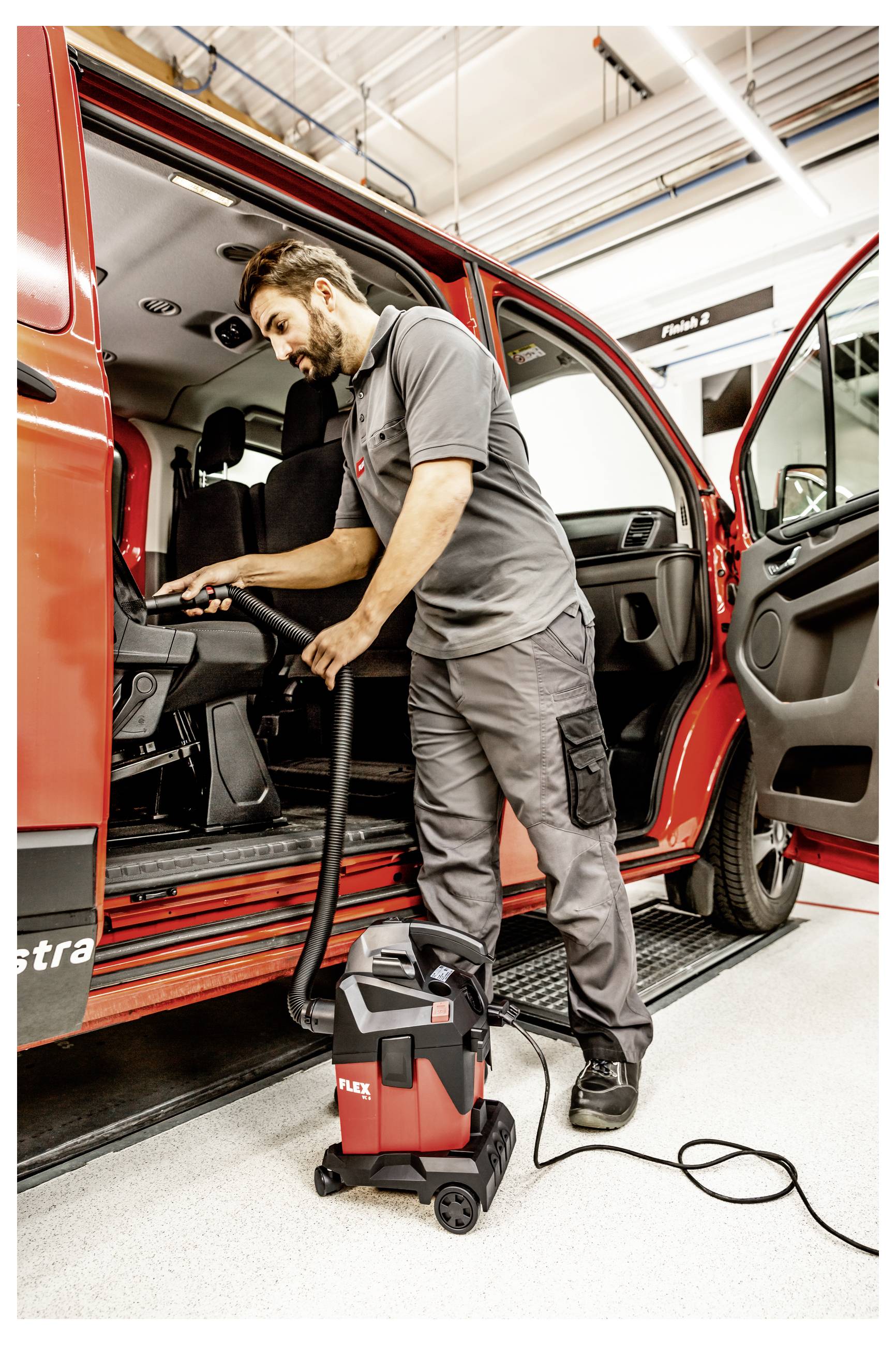 Een monteur die het interieur van een rode bestelwagen schoonmaakt met een draagbare stofzuiger in een werkplaats.