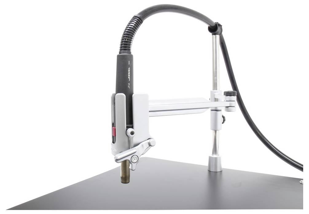 Close-up van een handheld warmtegereedschap bevestigd op een metalen arm boven een plat werkoppervlak, met een kabel eraan bevestigd.