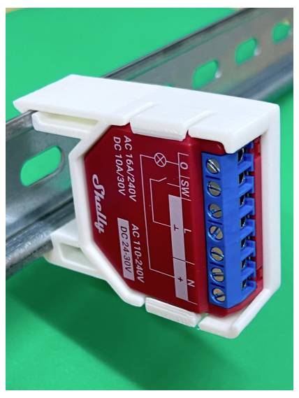 Close-up van een Shelly elektrische relaismodule gemonteerd op een DIN-rail, met blauwe schroefklemmen en gelabelde AC/DC-bedrading aansluitingen.