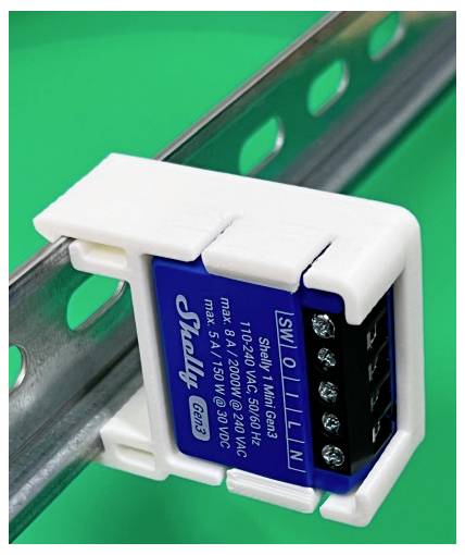 Close-up van een Shelly 1 Mini Gen3 slimme relaismodule gemonteerd op een metalen DIN-rail tegen een groene achtergrond.