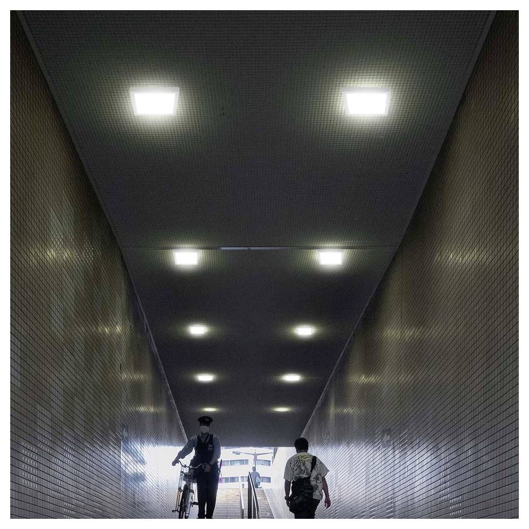 Twee personen lopen door een ondergrondse tegeldoorgang naar het daglicht, één met een fiets voortduwend, onder rijen plafondverlichting.