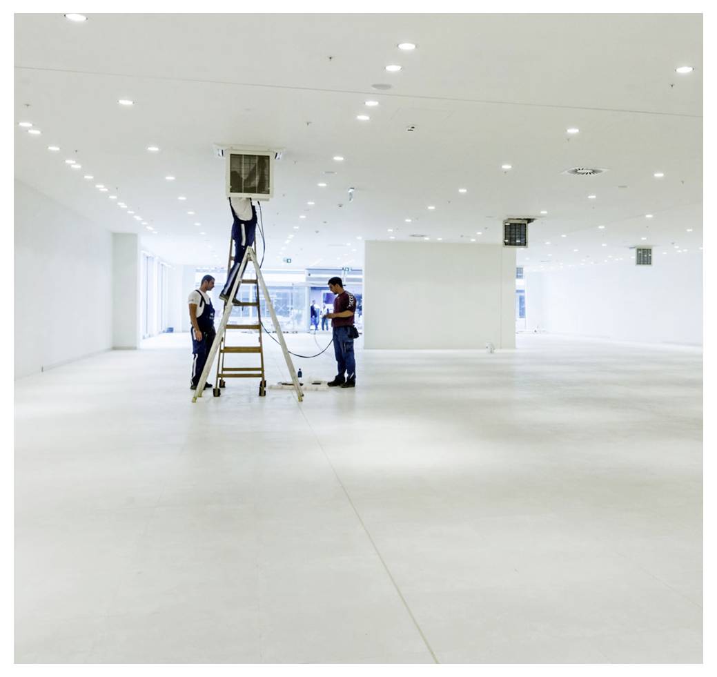 Arbeiders die een plafondventilatie in een groot, leeg wit commercieel interieur installeren met ladder en ingebouwde verlichting.