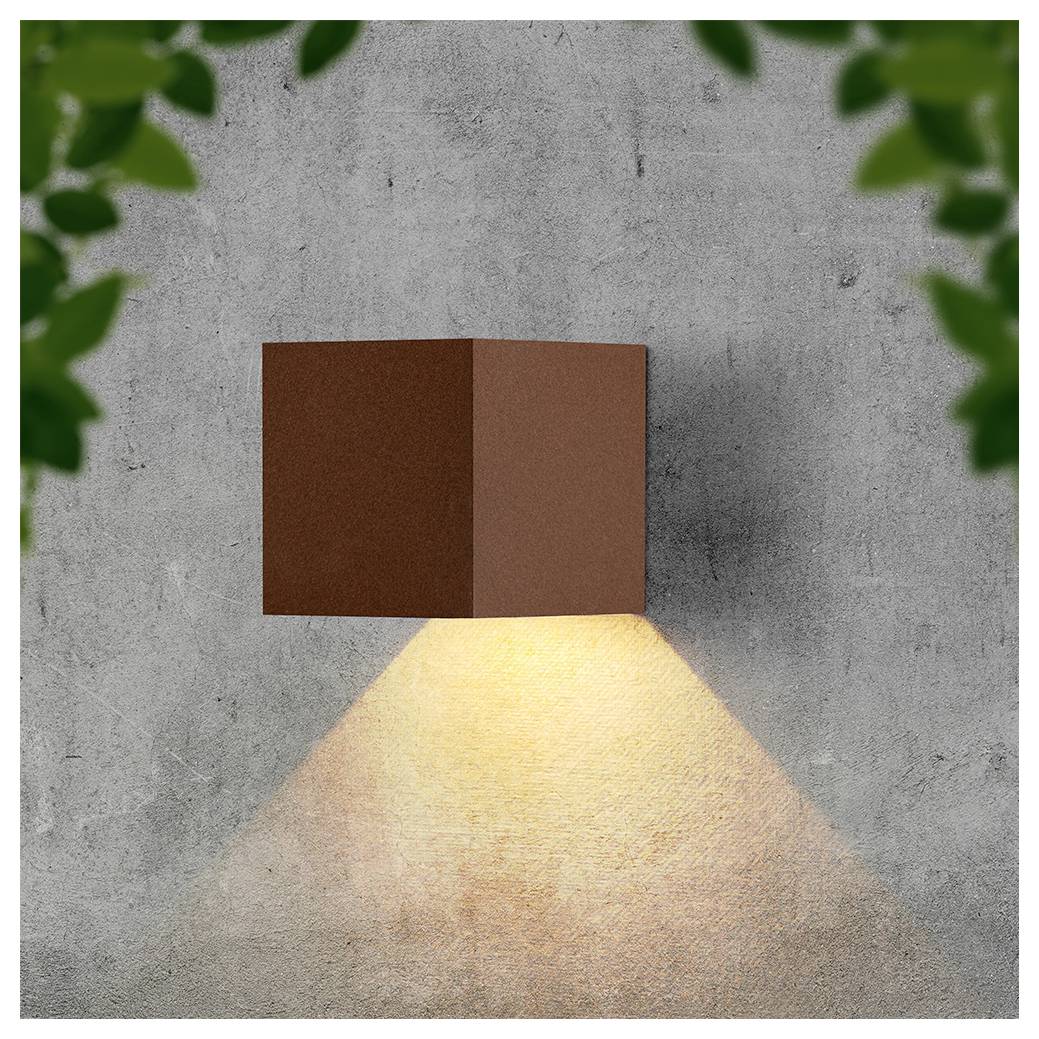 Aan de muur gemonteerde bruine kubuslamp werpt een warm driehoekig lichtstraal naar beneden op een grijze betonnen muur, omlijst door vervaagde groene bladeren.