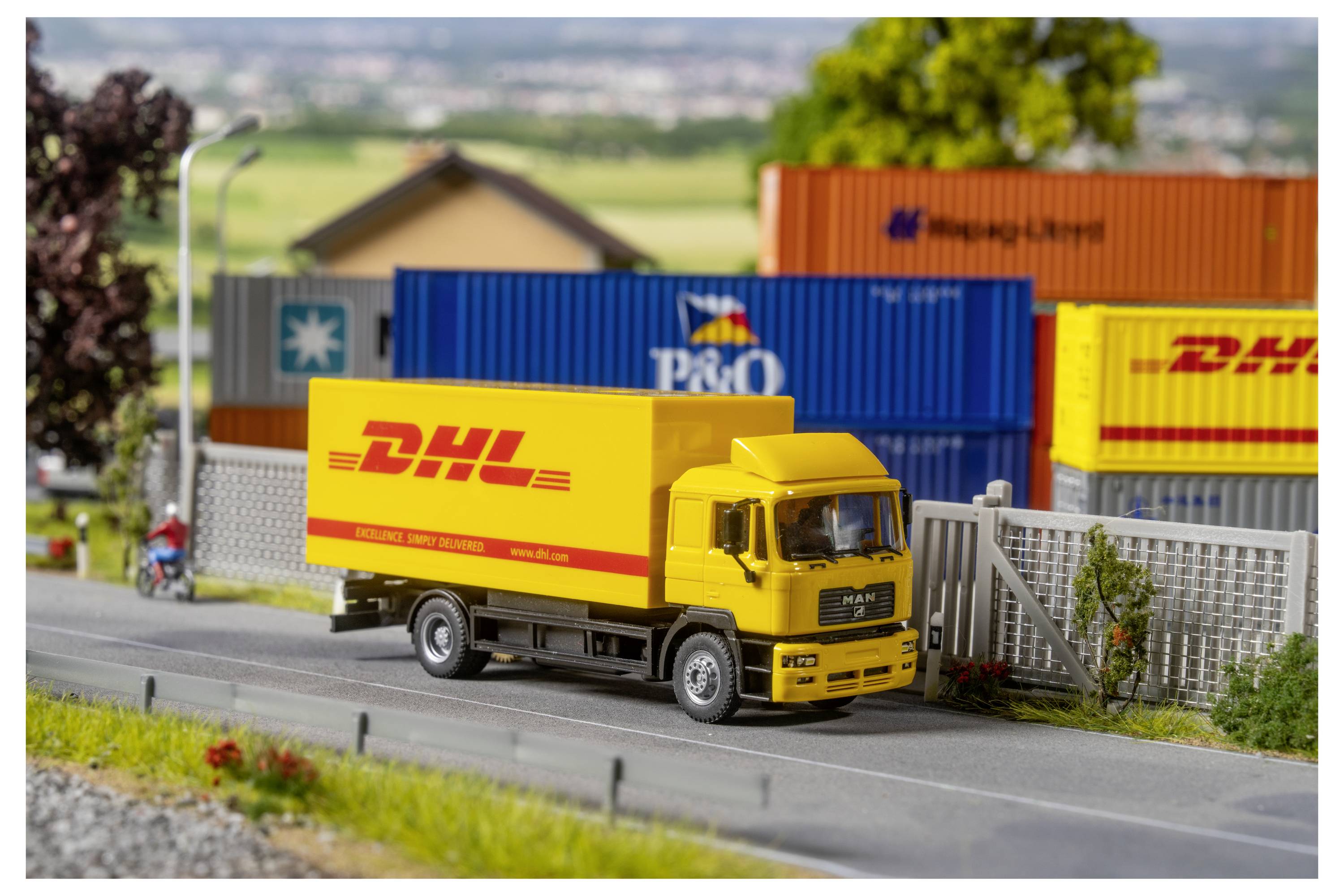 Gele DHL-bezorgingsvrachtwagen die op een weg rijdt naast gestapelde scheepscontainers in een miniatuurtafereel op een vrachtterrein.