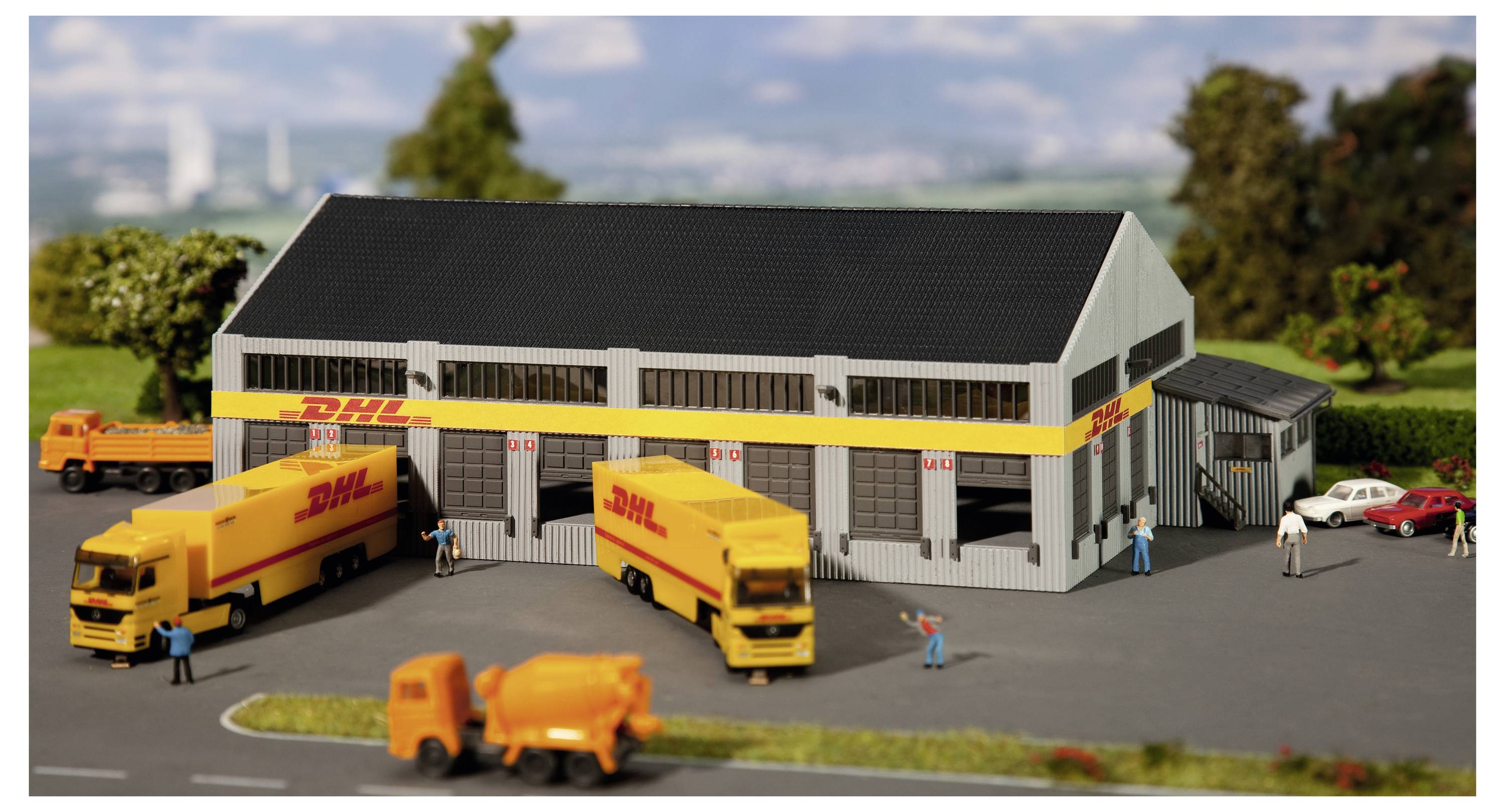 Miniatuurdiorama van DHL-logistiekdepot met meerdere gele bezorgvrachtwagens, werknemers, een tankwagen en geparkeerde auto's buiten een magazijngebouw.