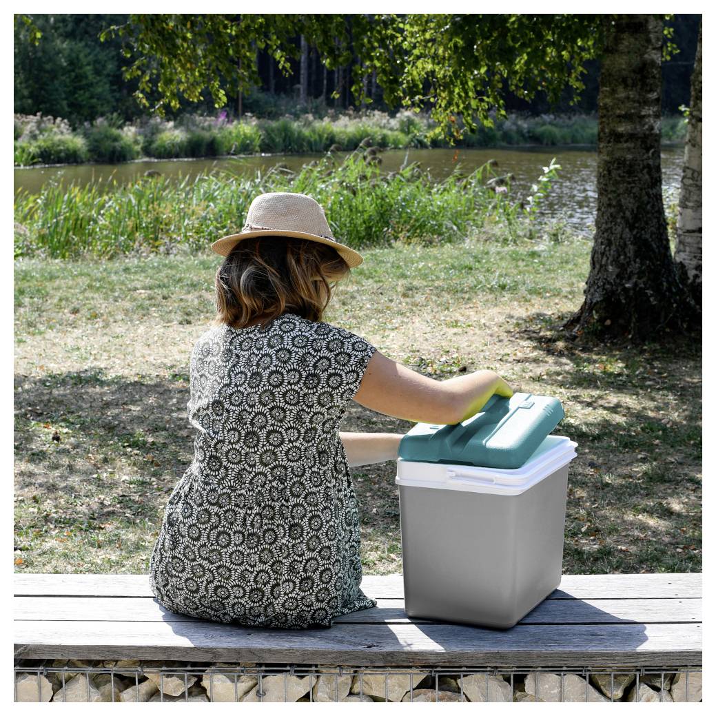 Een vrouw met een hoed zit op een bank bij een vijver en opent een koelbox of opslagdoos buiten in een park.