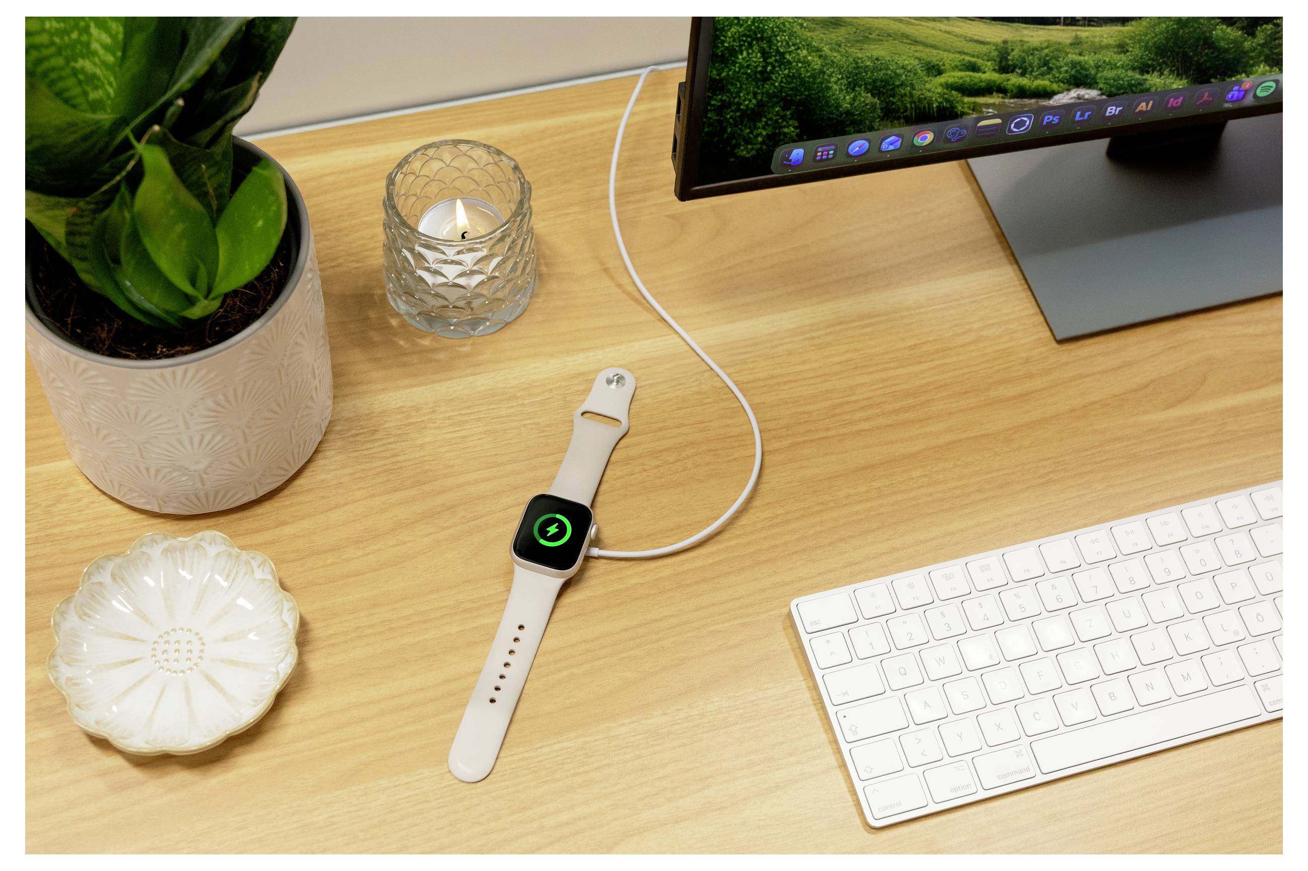 Apple Watch met een wit bandje dat oplaadt op een houten bureau naast een toetsenbord, monitor, plant en kaarsen.