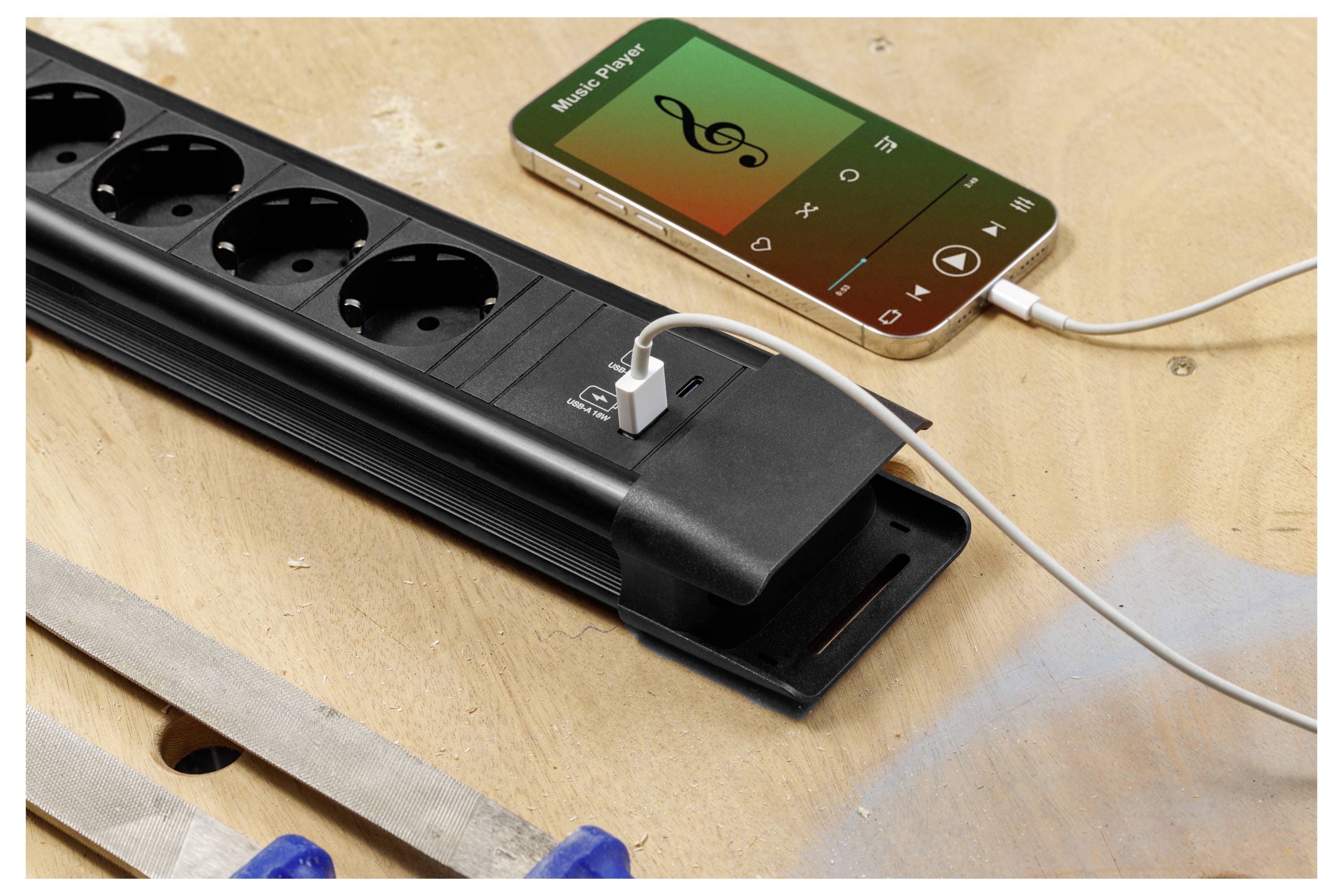 Een smartphone speelt muziek af terwijl deze naast een zwart stroomverdeelblok met meerdere stopcontacten en USB-poorten op een werkbank wordt opgeladen.