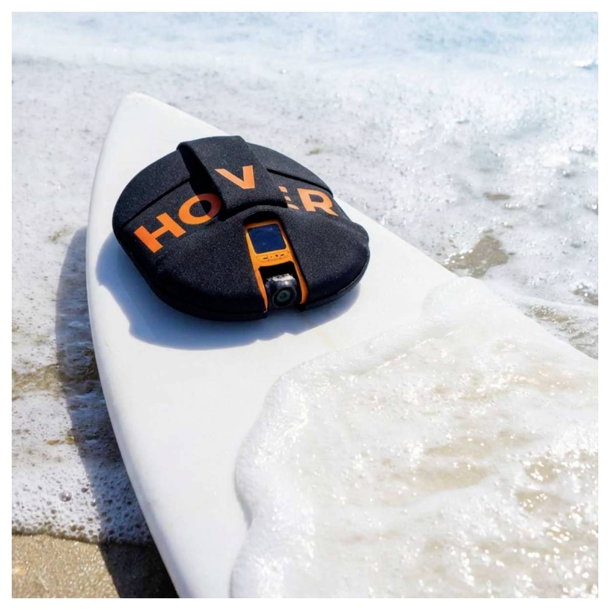 Zwart en oranje 'HOVER' apparaat bevestigd aan een witte surfboard op een schuimende golf aan het strand