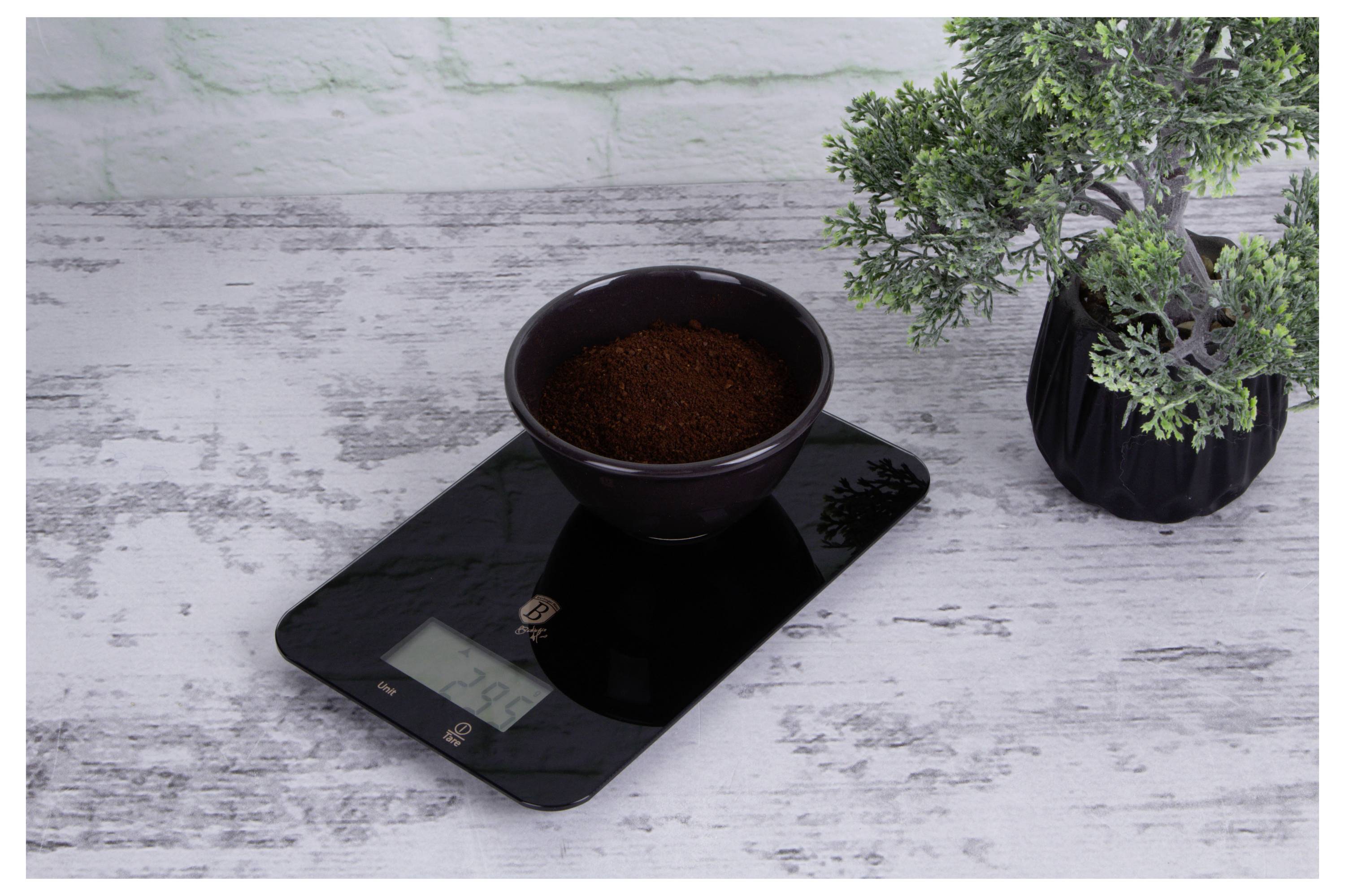 Koffiedik in een kom op een digitale keukenweegschaal naast een klein potje plant op een wit houten oppervlak.
