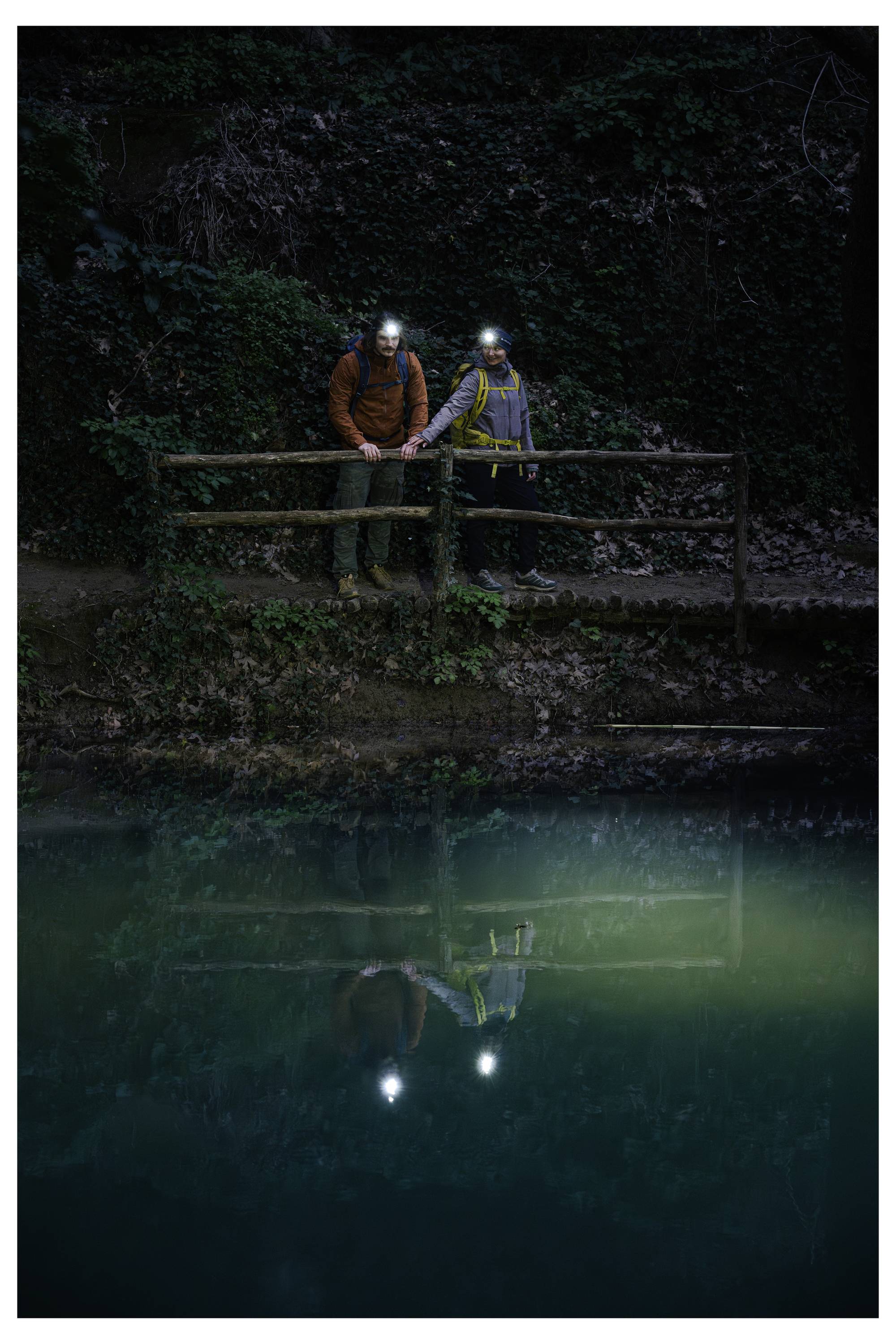Twee wandelaars met koplampen staan op een houten brug boven donker water, met hun weerspiegeling zichtbaar eronder.