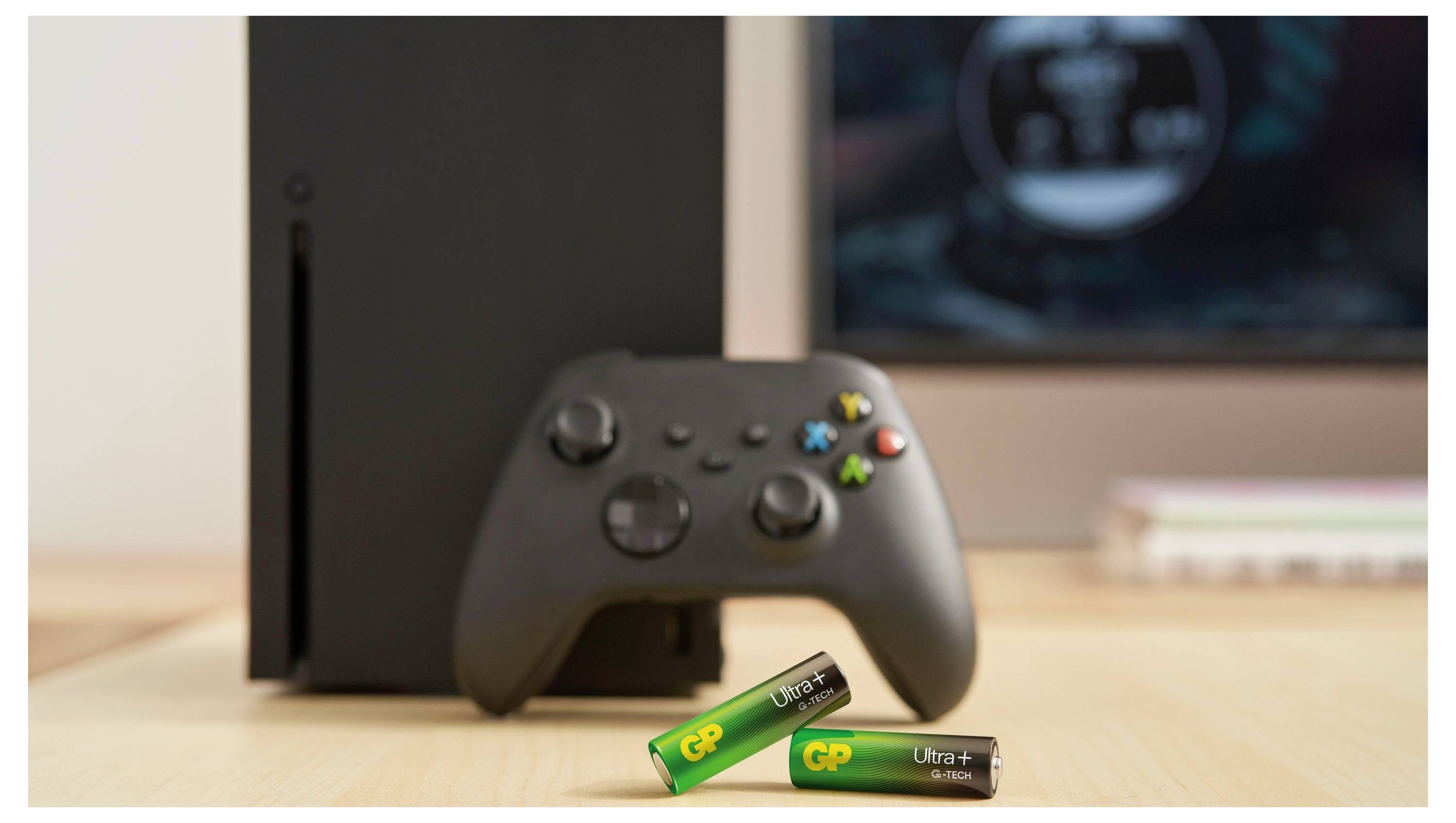 Zwart Xbox-console met controller en twee GP Ultra AA-batterijen op een bureau voor een monitor.