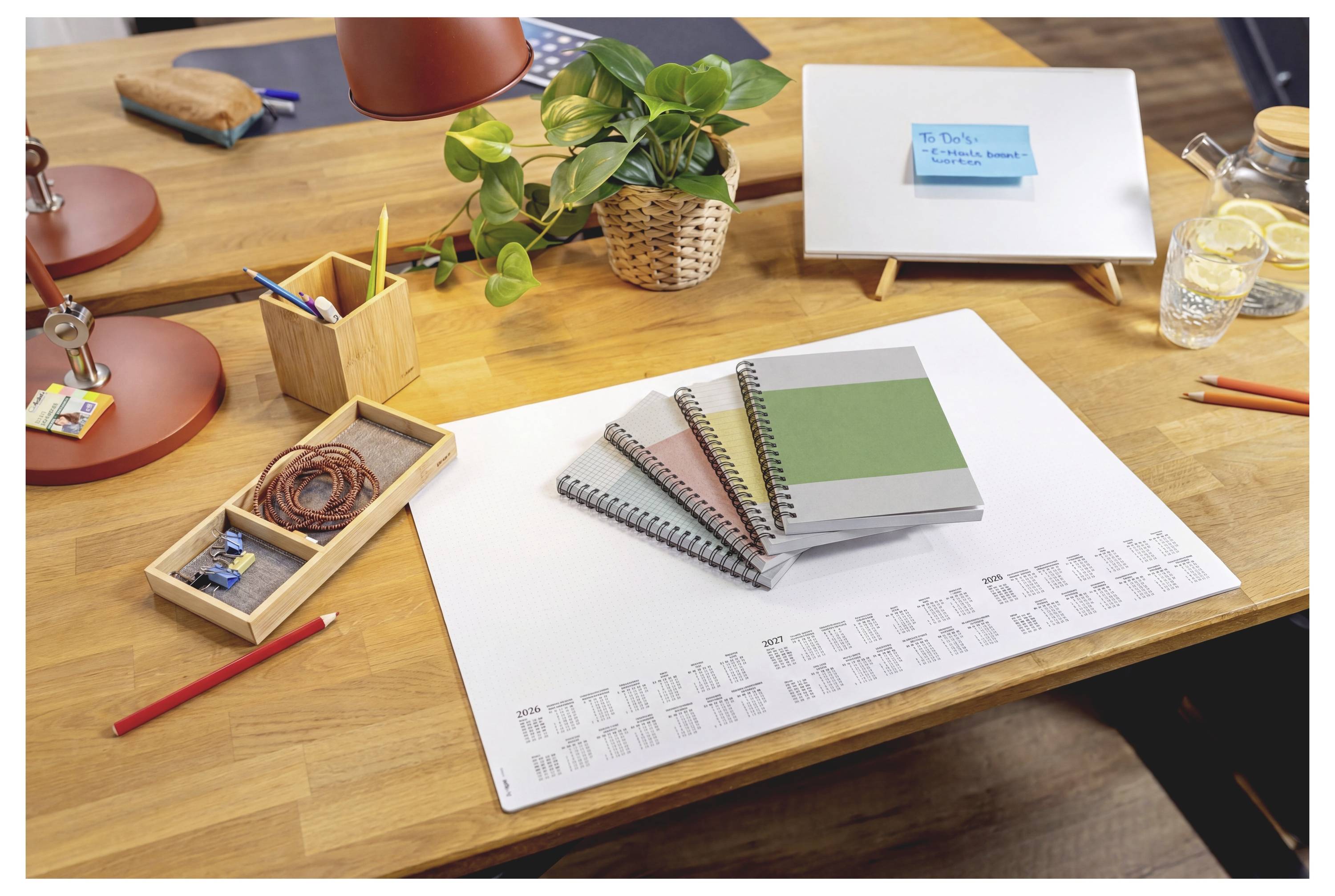 Persoon die schrijft in een notitieboek aan een bureau met een klein whiteboard met een blauwe kleefnota, een plant en een kan citroenwater in de buurt.