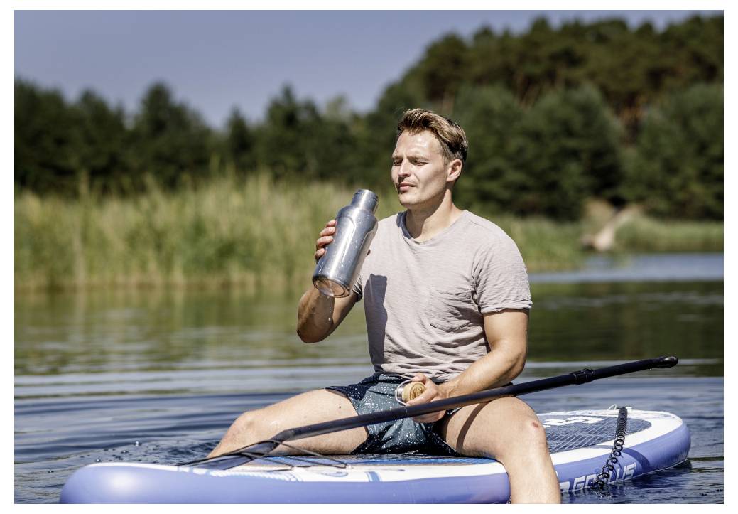 Een man zit op een peddelbord op een meer, drinkt uit een metalen waterfles terwijl hij een peddel vasthoudt.