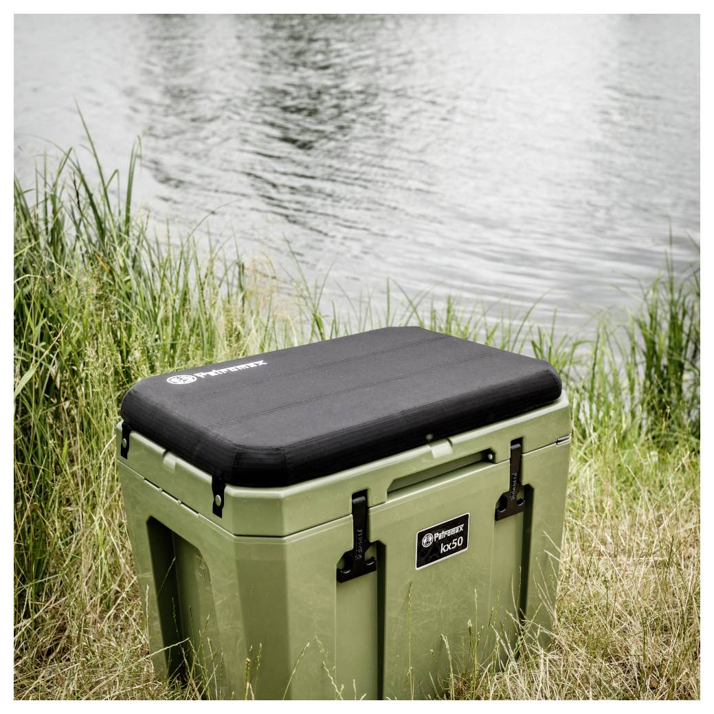Olijfgroene draagbare koelbox met een zwart deksel staat in het hoge gras naast een meer.