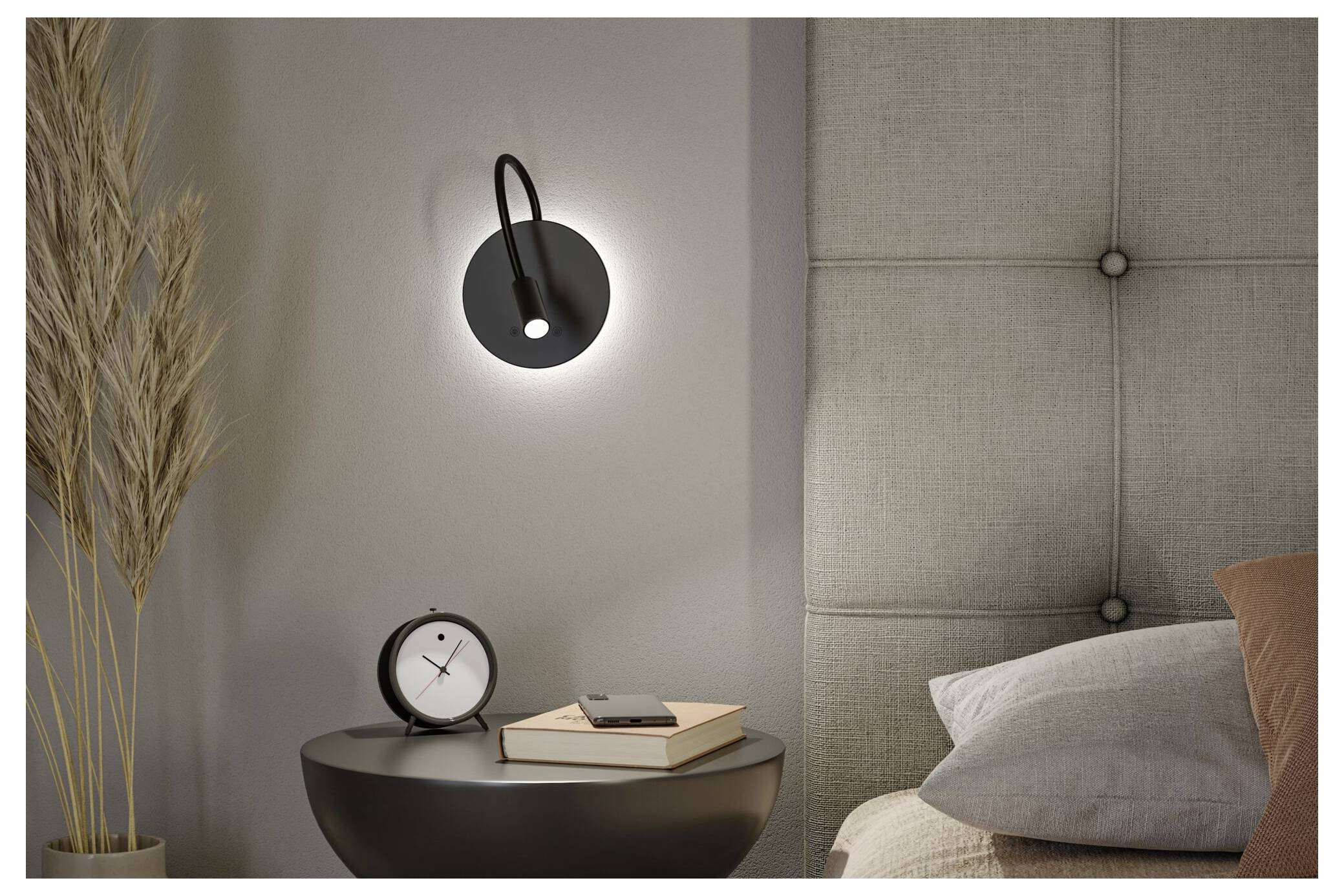 Slaapkamertafereel naast het bed met een aan de muur gemonteerde zwarte lamp, klein rond tafeltje, boeken, klok, droge grassen en kussens tegen een gestoffeerd hoofdeinde.