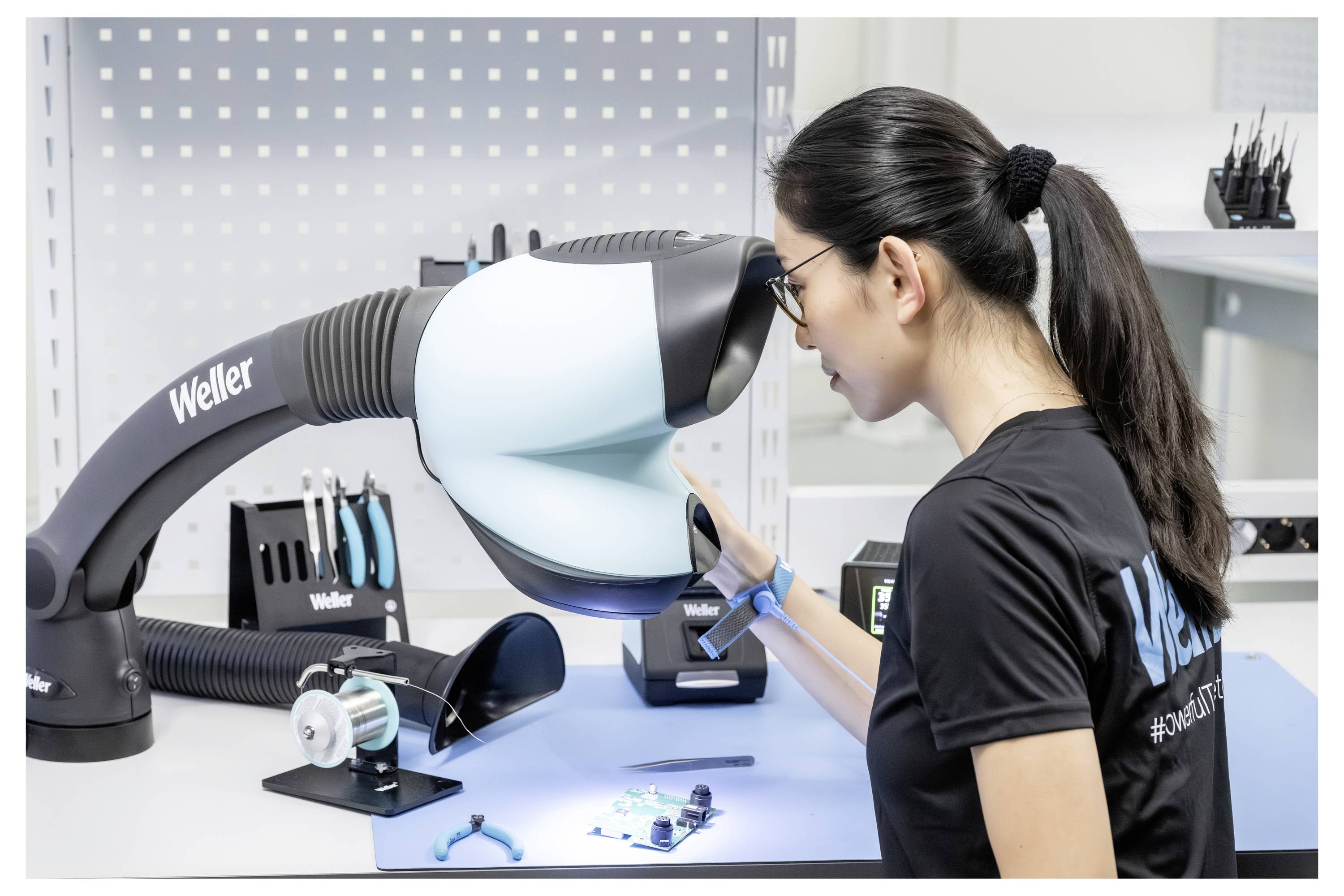 Vrouw in een zwart shirt inspecteert een robotisch werkbanktoestel met het label Weller in een schoon elektronicalab.