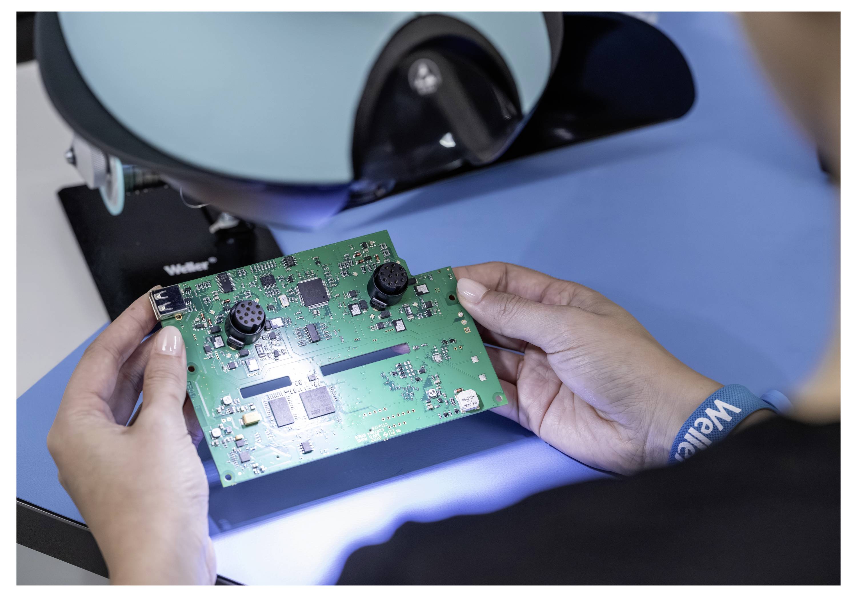 Handen die een groen printplaatje onder een inspectie-microscoop houden, wat duidt op kwaliteitscontrole of reparatie van elektronische apparatuur.