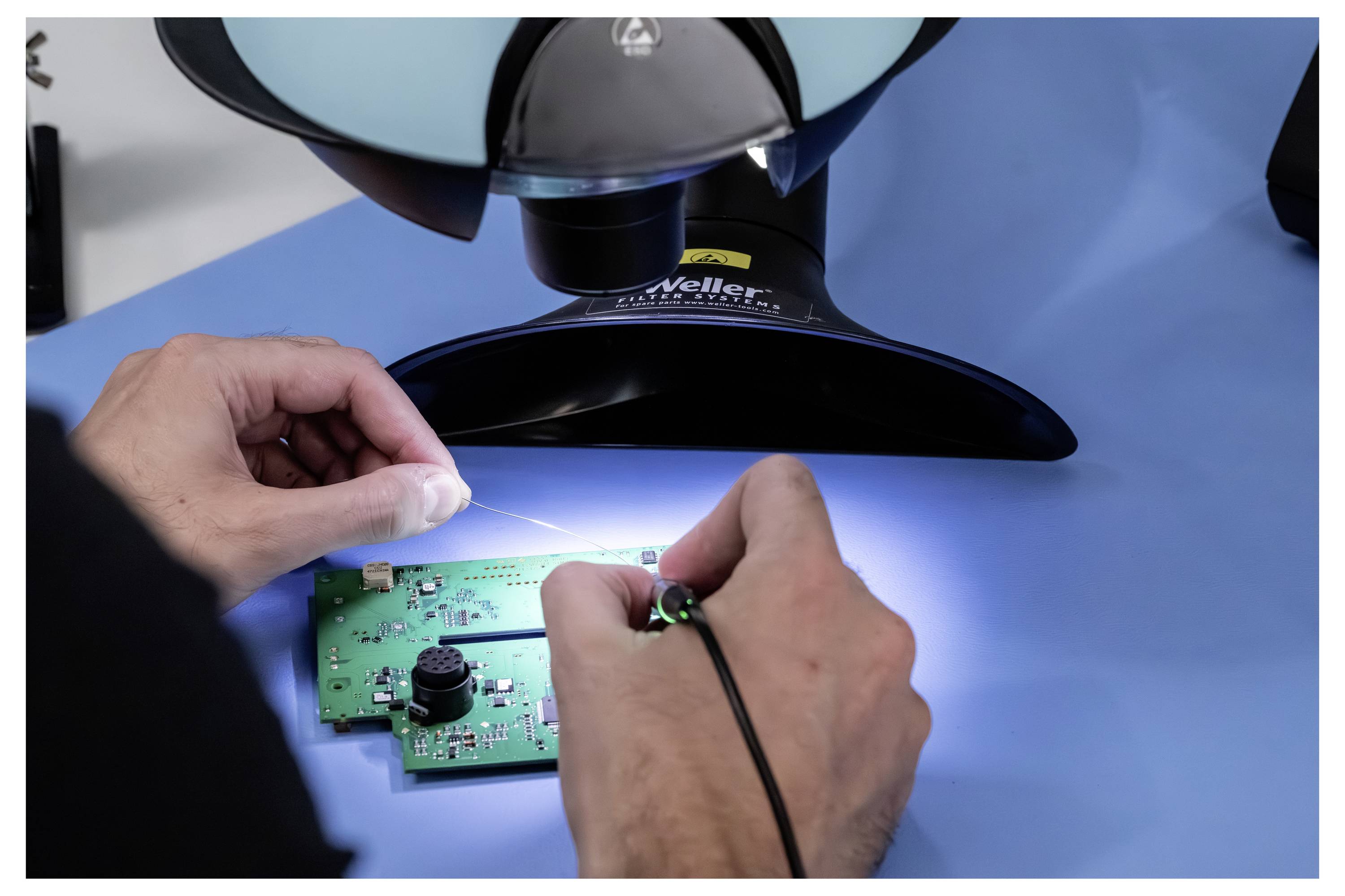 Handen solderen een klein groen printplaat onder een microscoop op een blauw werkoppervlak, wat wijst op elektronicarepratie of inspectie.
